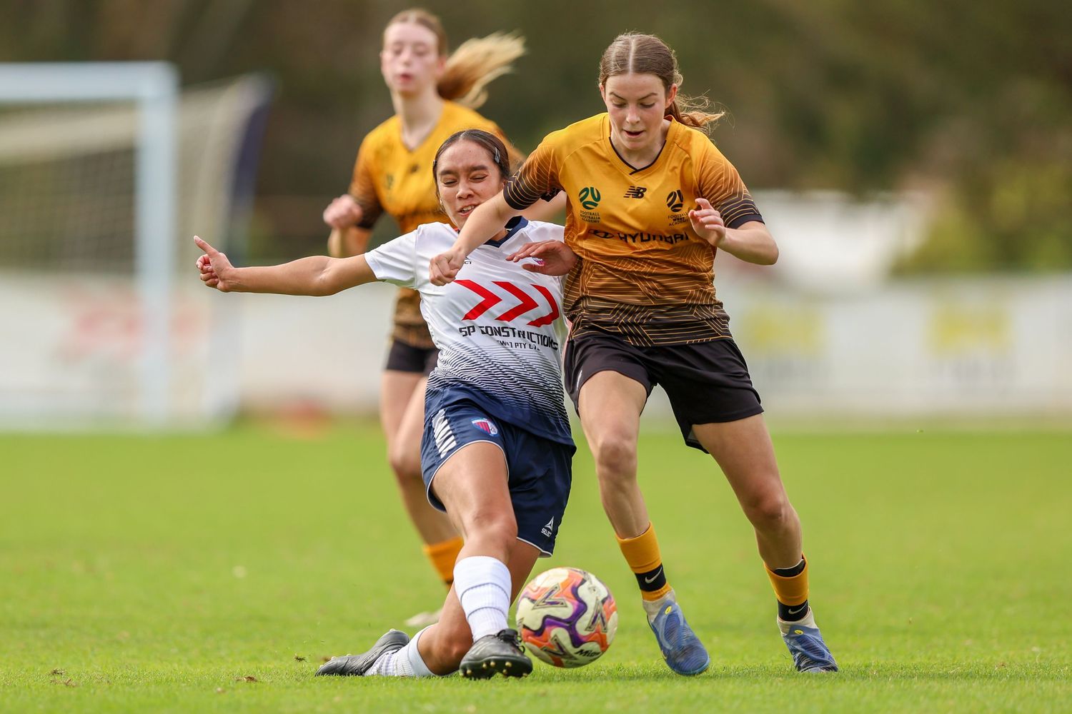 Two female soccer players compete for the ball during a youth football match on a green pitch.