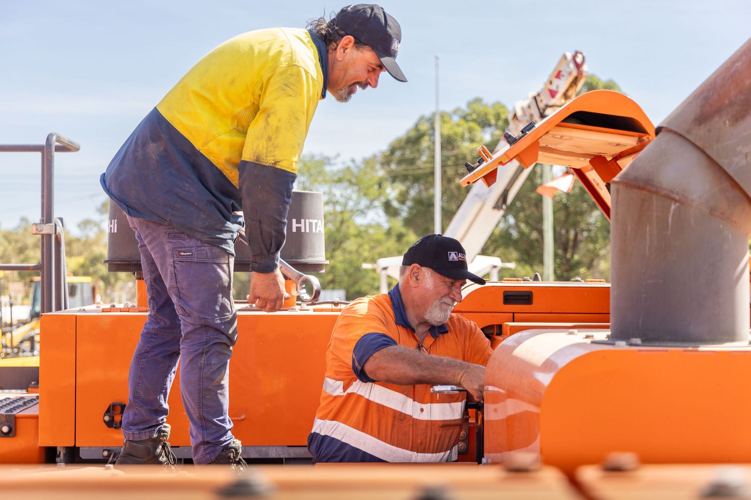 Construction workers in hard hats operate orange industrial equipment on a sunny worksite.
