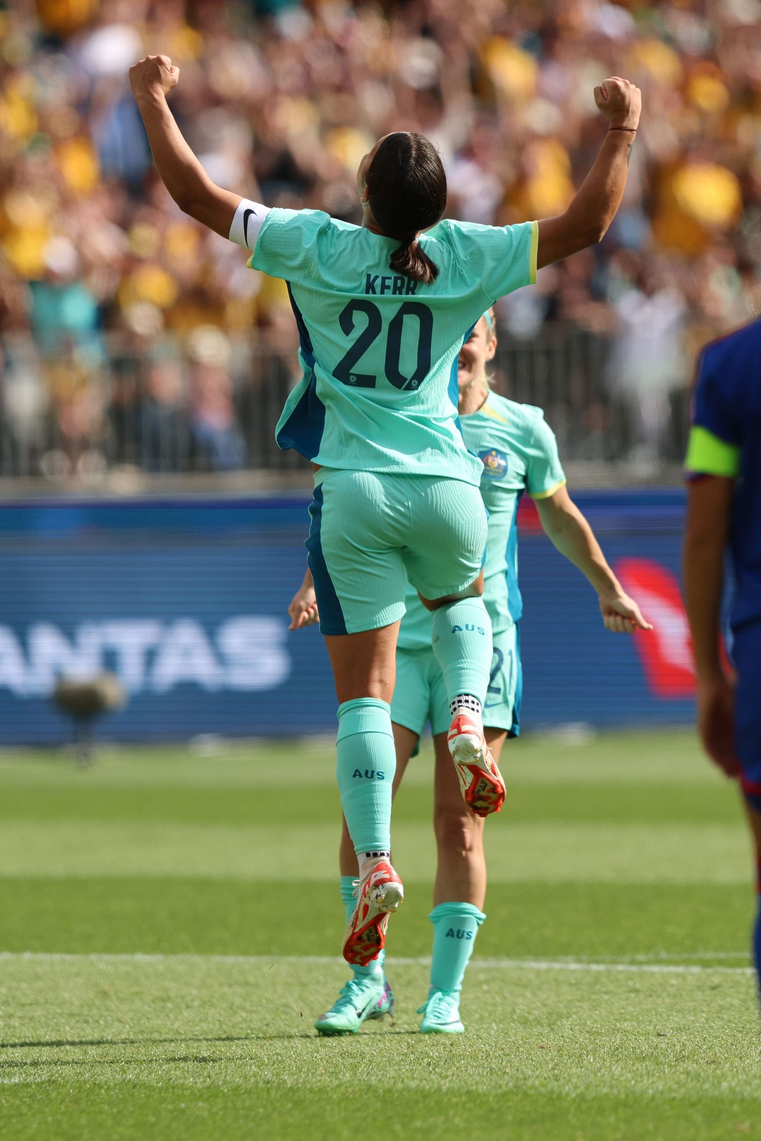 Soccer player in turquoise jersey number 20 celebrates with raised arms on a sunny stadium field.