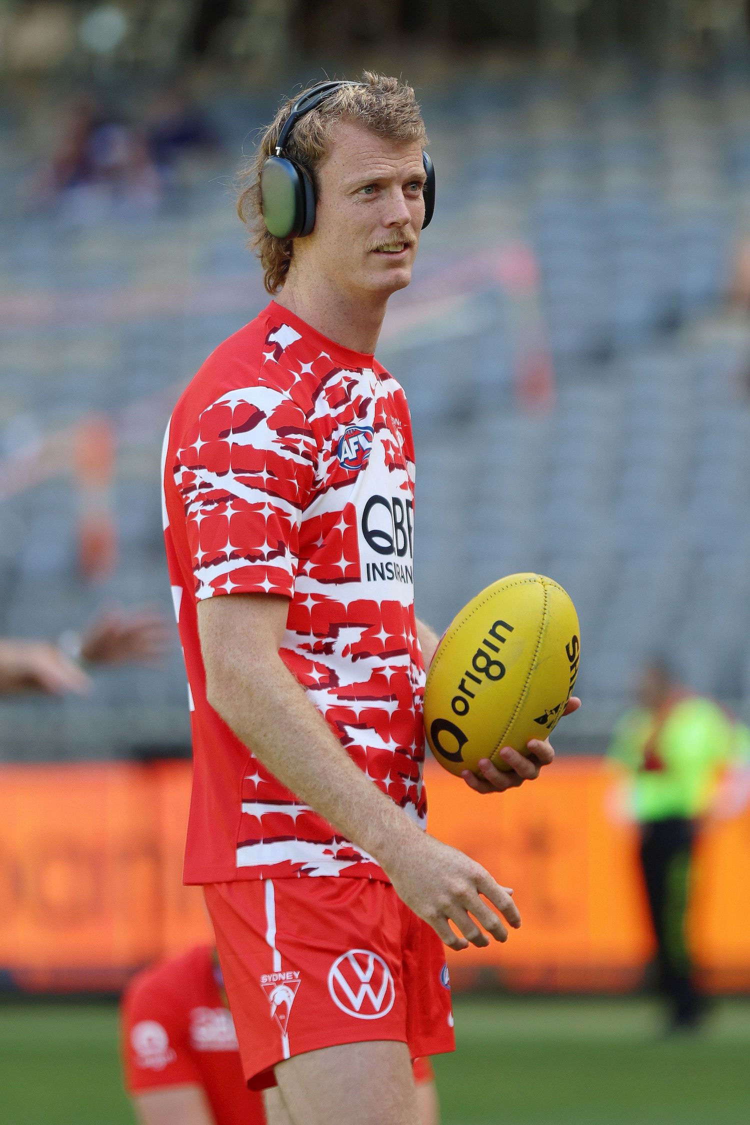 Player in red patterned uniform wearing headphones holds yellow football on field.