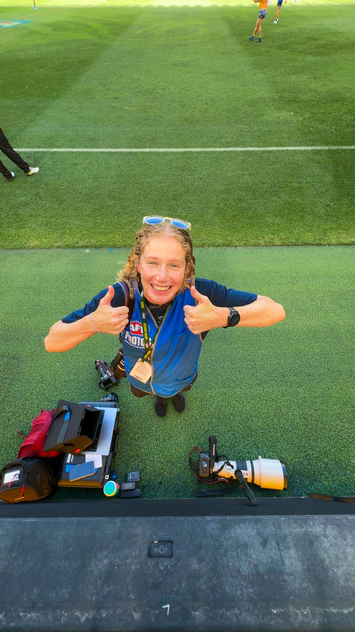 Sports photographer with camera equipment giving thumbs up on field sideline.