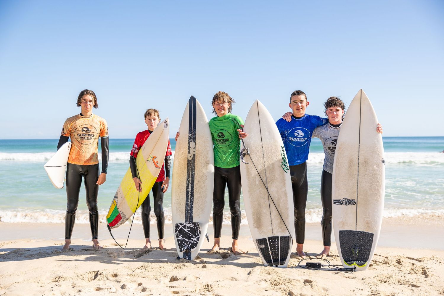 Surfers lined up on the beach with their surfboards against a bright blue ocean and sky background.