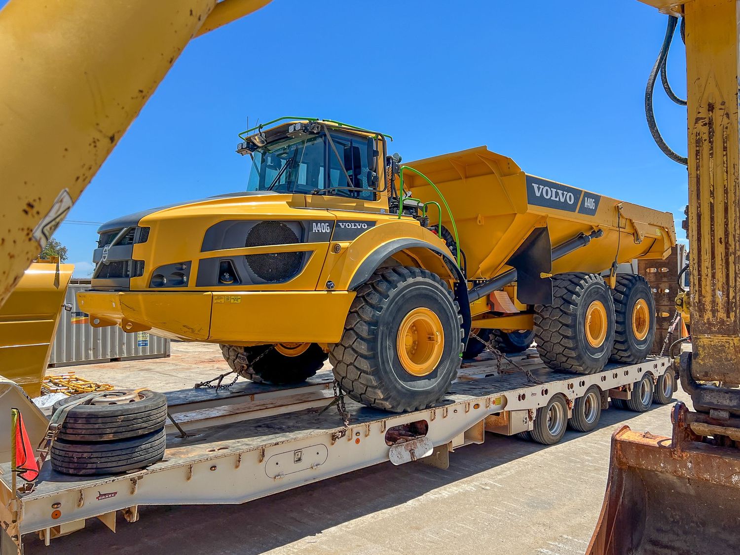 Large yellow articulated dump truck being transported on a flatbed trailer in bright sunlight.