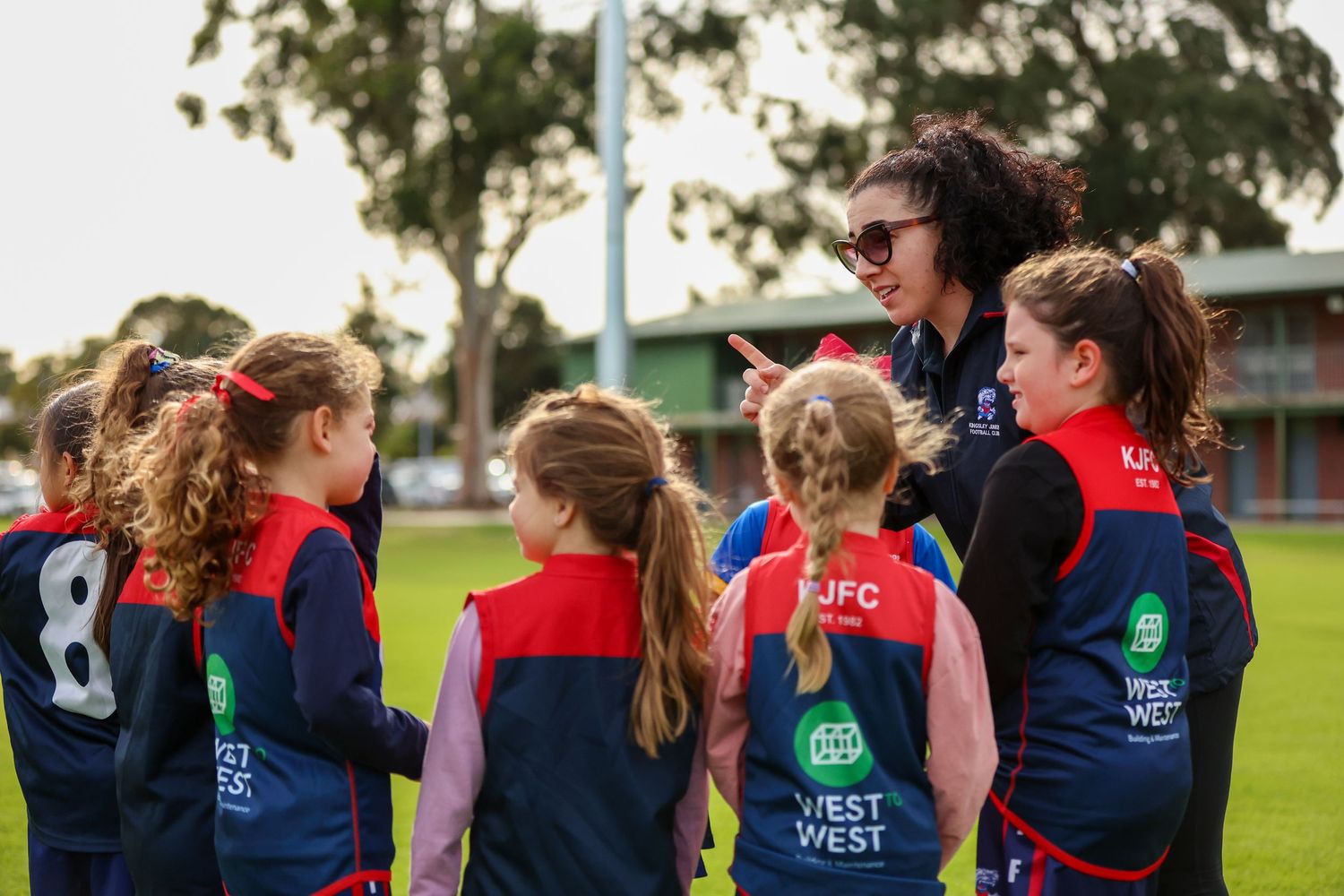 A youth sports coach and players in navy and red uniforms gather on a sports field during practice.