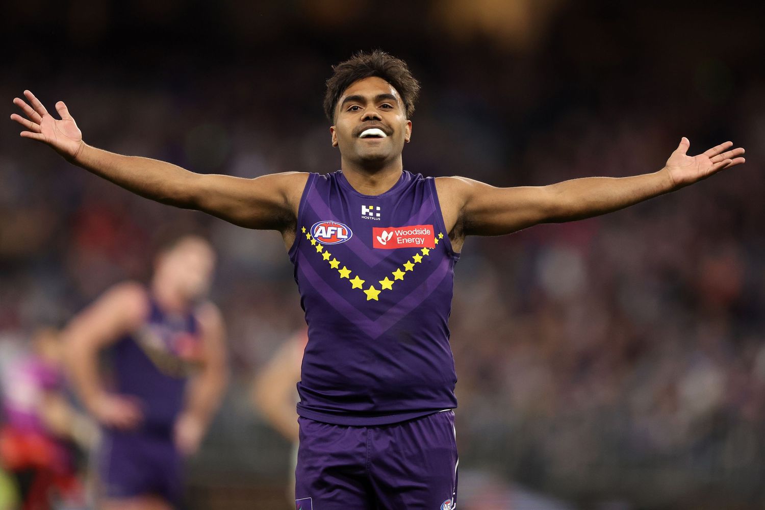 AFL players in purple jerseys celebrate with arms raised during an exciting match moment at a crowded stadium.