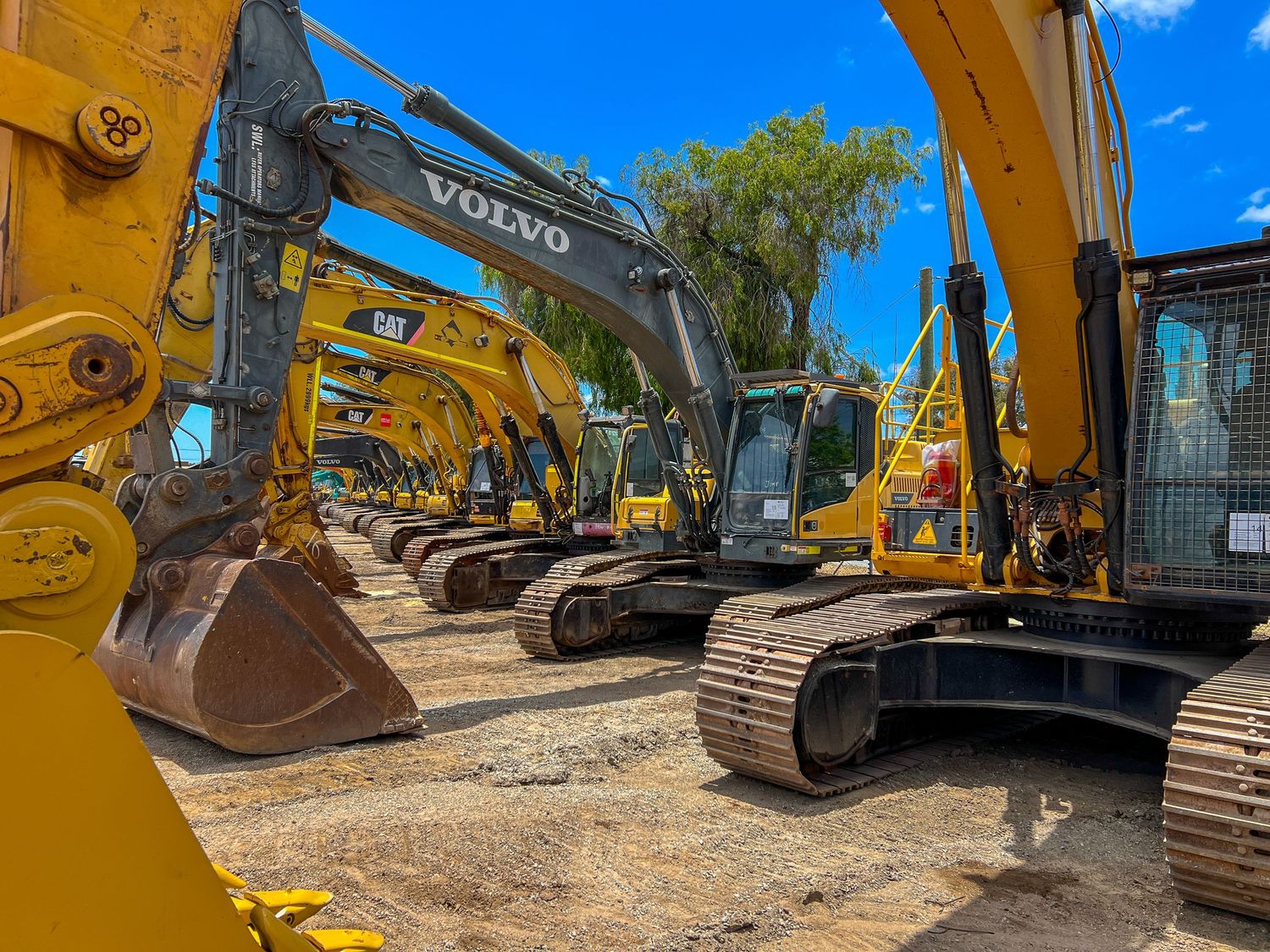 Row of yellow and gray excavators and construction equipment lined up at an outdoor machinery yard under blue sky.