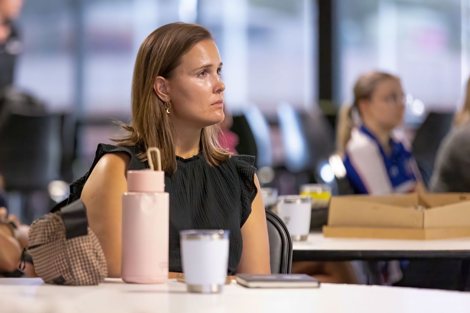 A person sits at a desk with a reusable water bottle and coffee cup in an office or classroom setting.