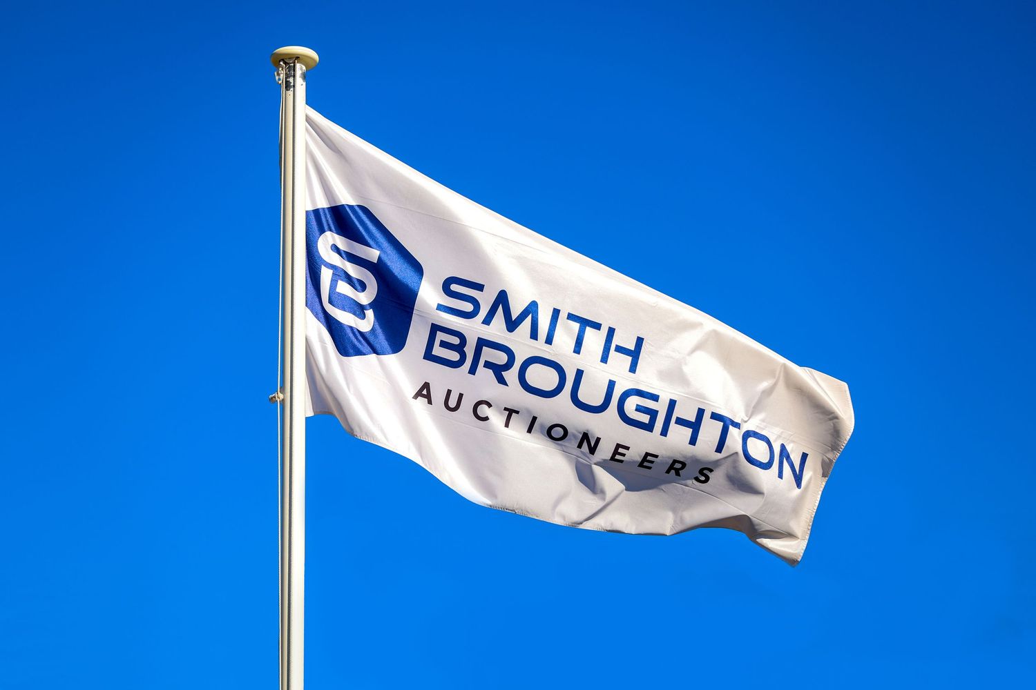 Smith Broughton Auctions white flag with blue logo waving against bright blue sky.