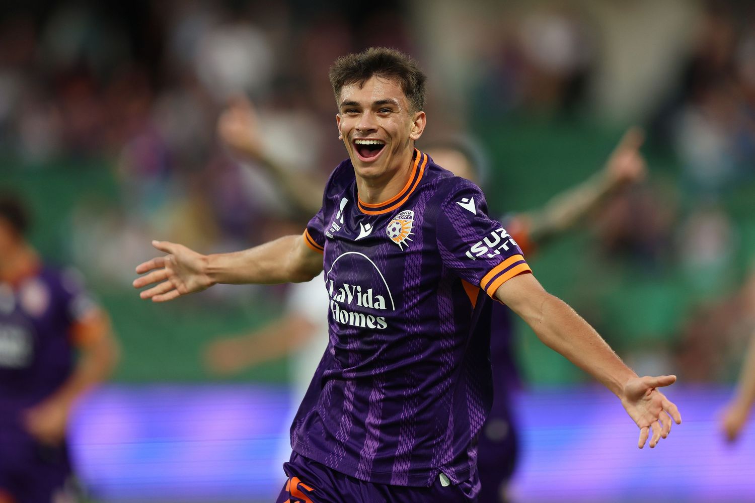 Soccer player celebrates in a purple jersey with arms outstretched during a match at a stadium.