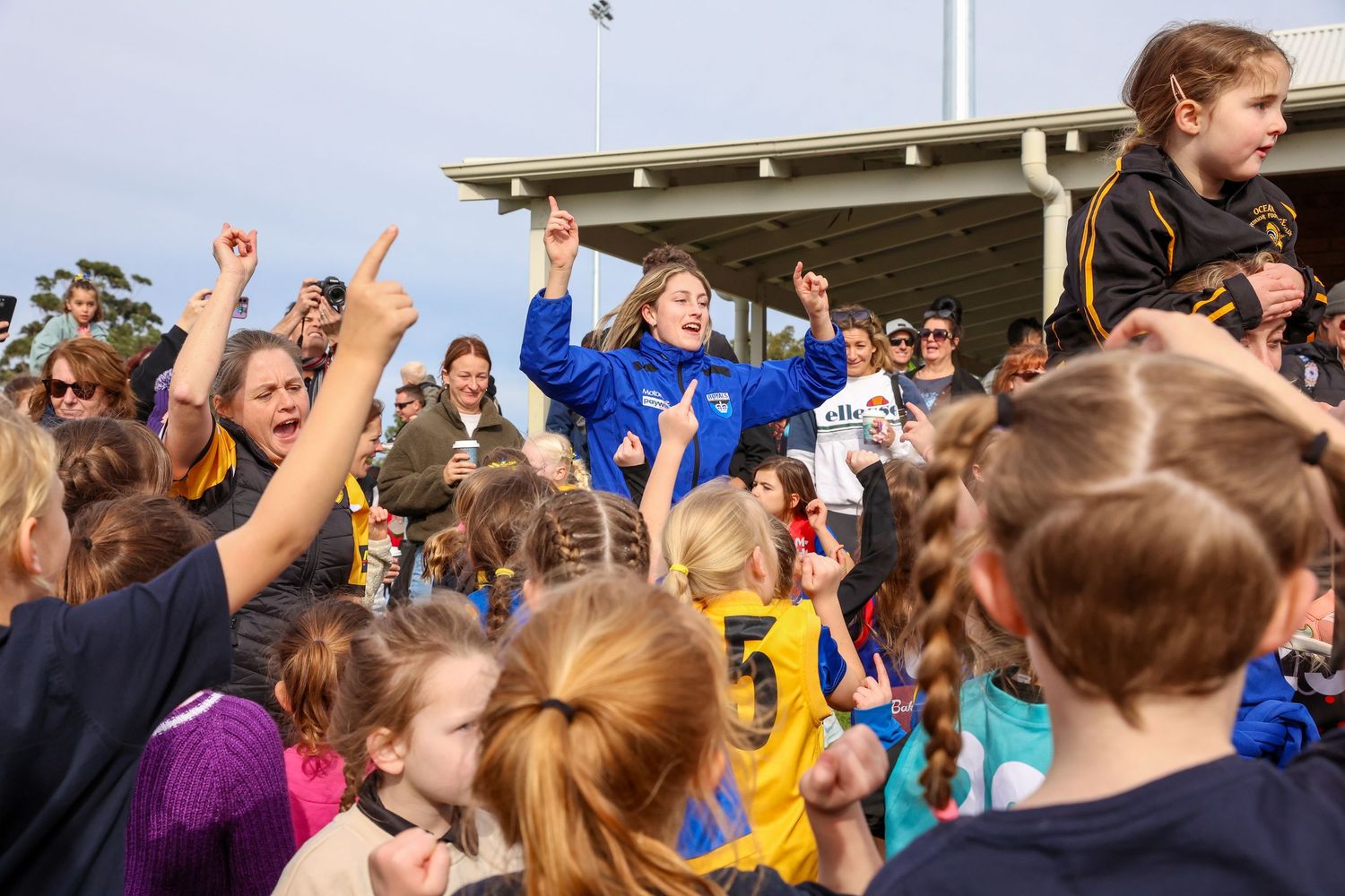 An excited crowd of students cheers and raises their hands during an outdoor school event.