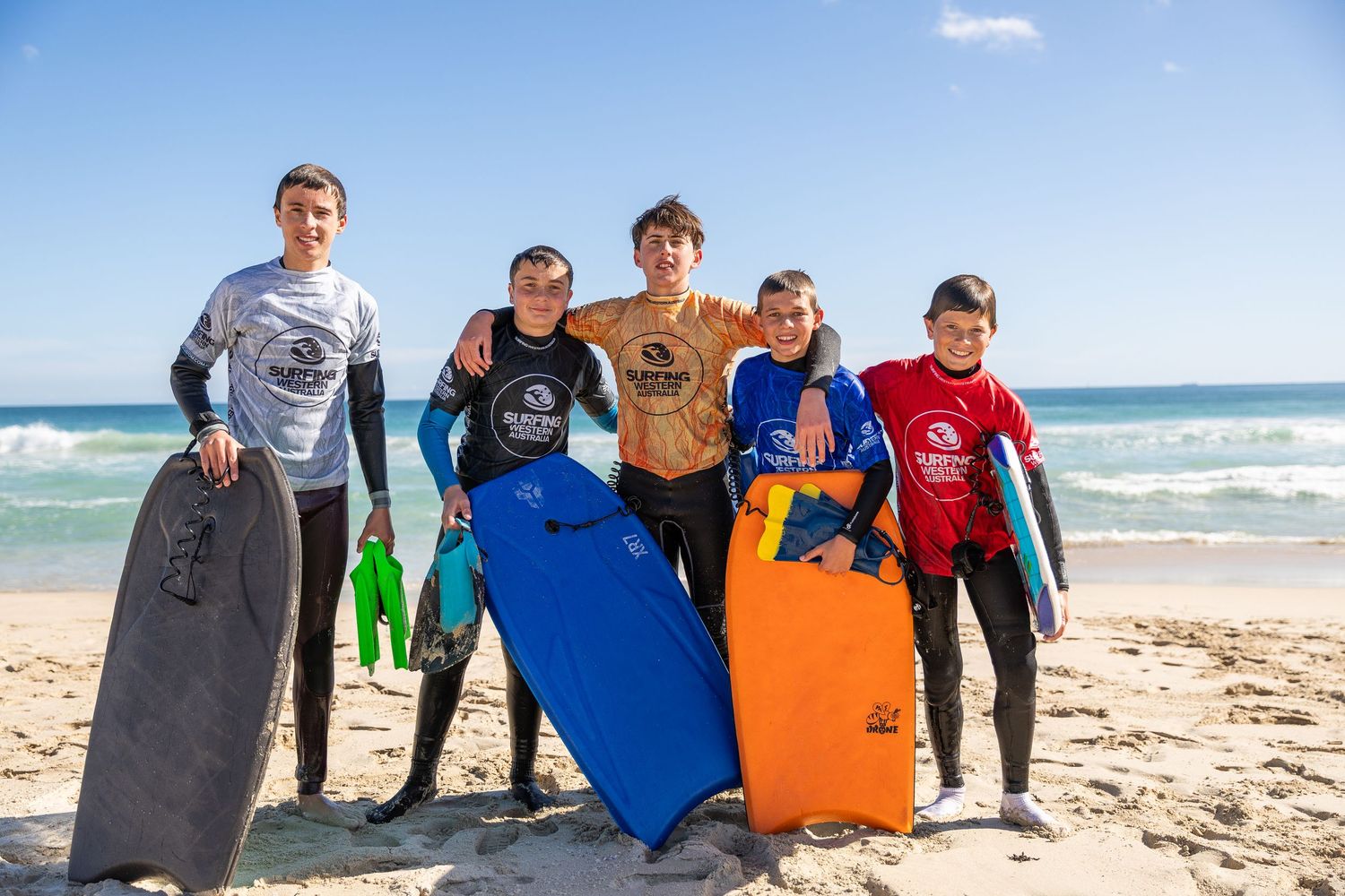 Group of surfers with bodyboards standing together on a sandy beach with ocean waves in the background.