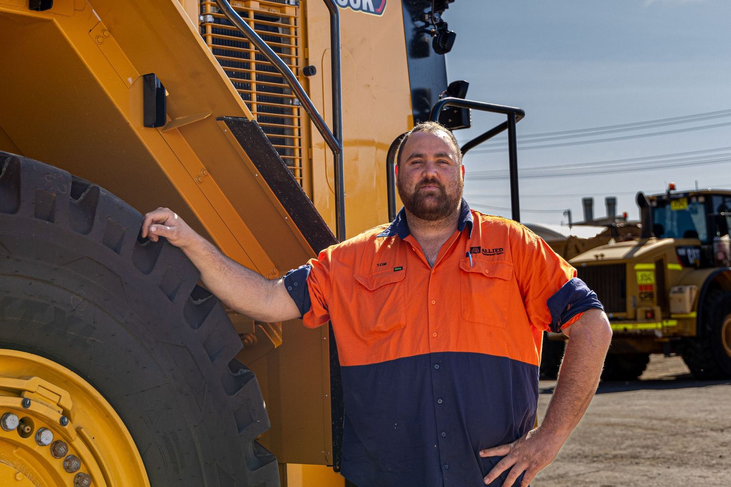 Construction worker in hi-vis orange and navy workwear stands confidently next to large yellow industrial equipment.