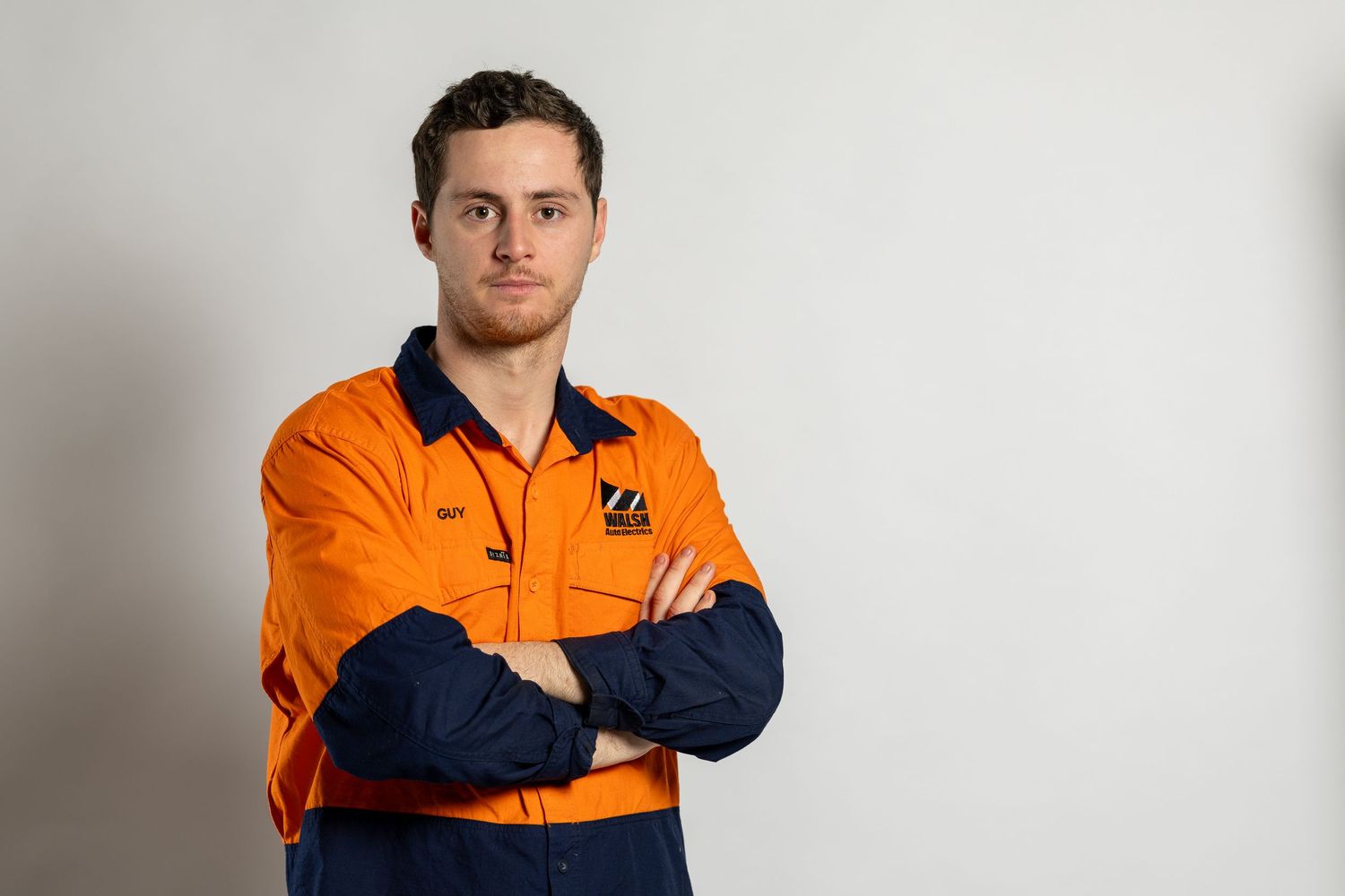 Worker in orange and navy hi-vis workwear stands with arms crossed against white background.