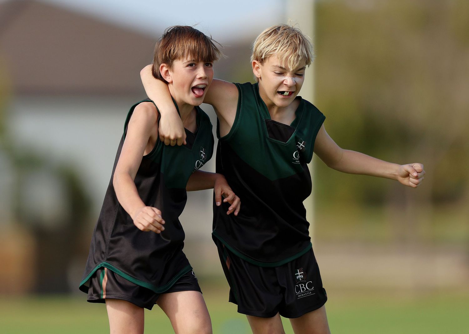 Two young athletes in dark green uniforms celebrate enthusiastically during an outdoor sports match.