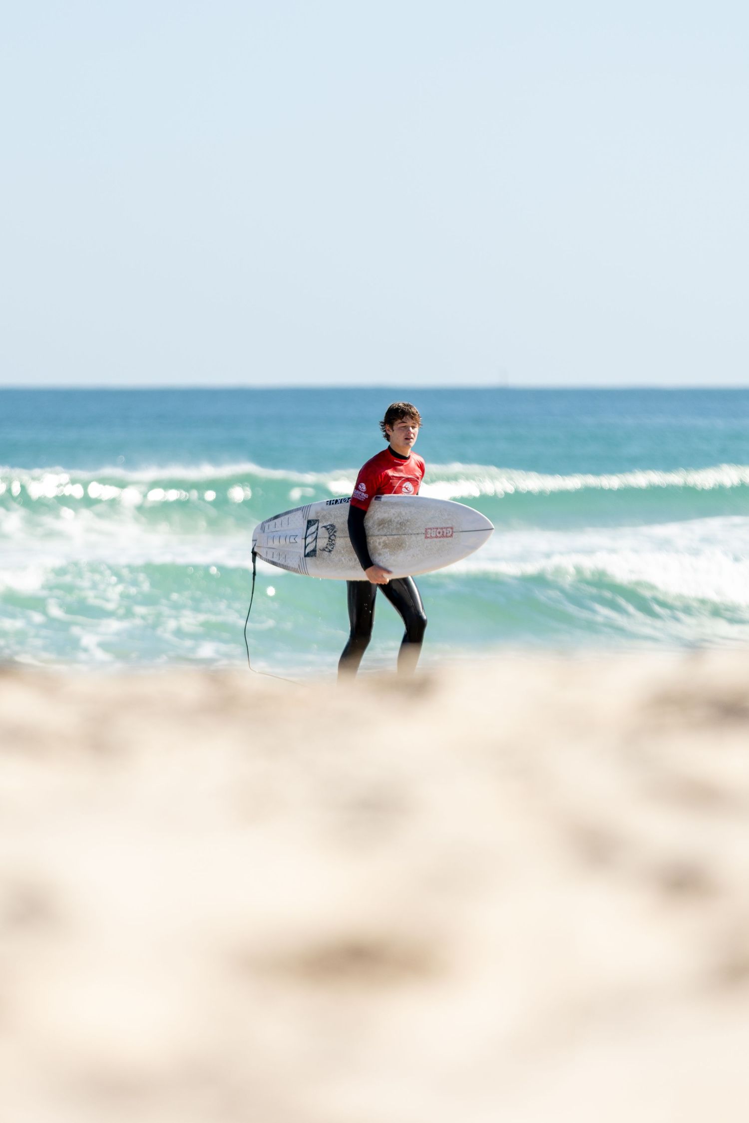 A surfer walks along the beach towards ocean waves carrying their surfboard on a sunny day.