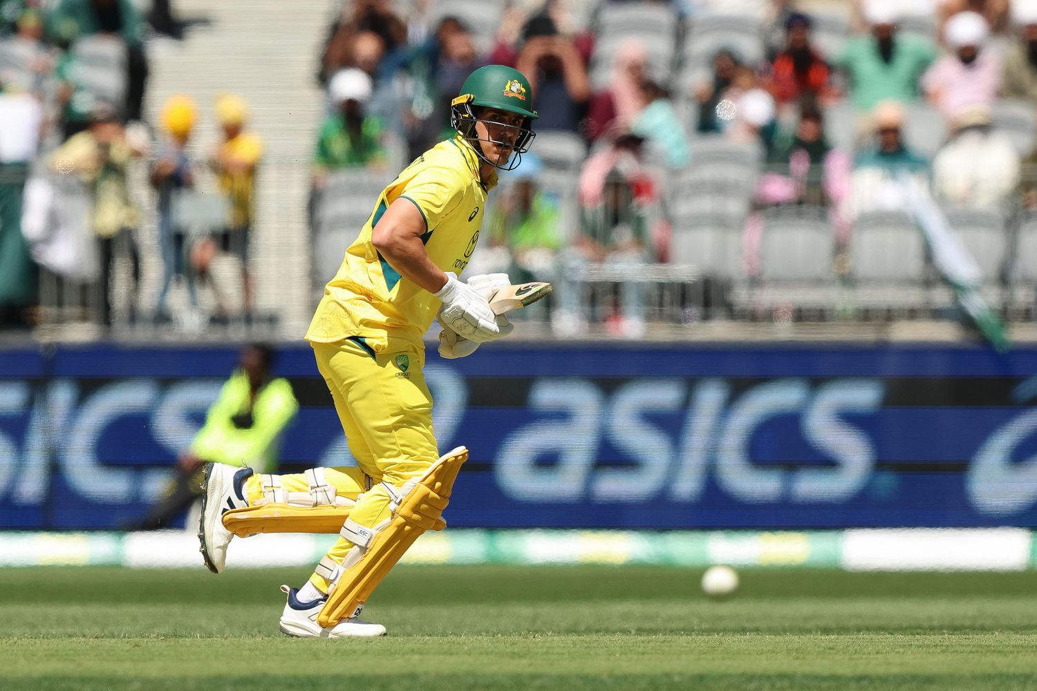 Cricket player in yellow Australian uniform runs between wickets during a match with Asics advertising visible in background.