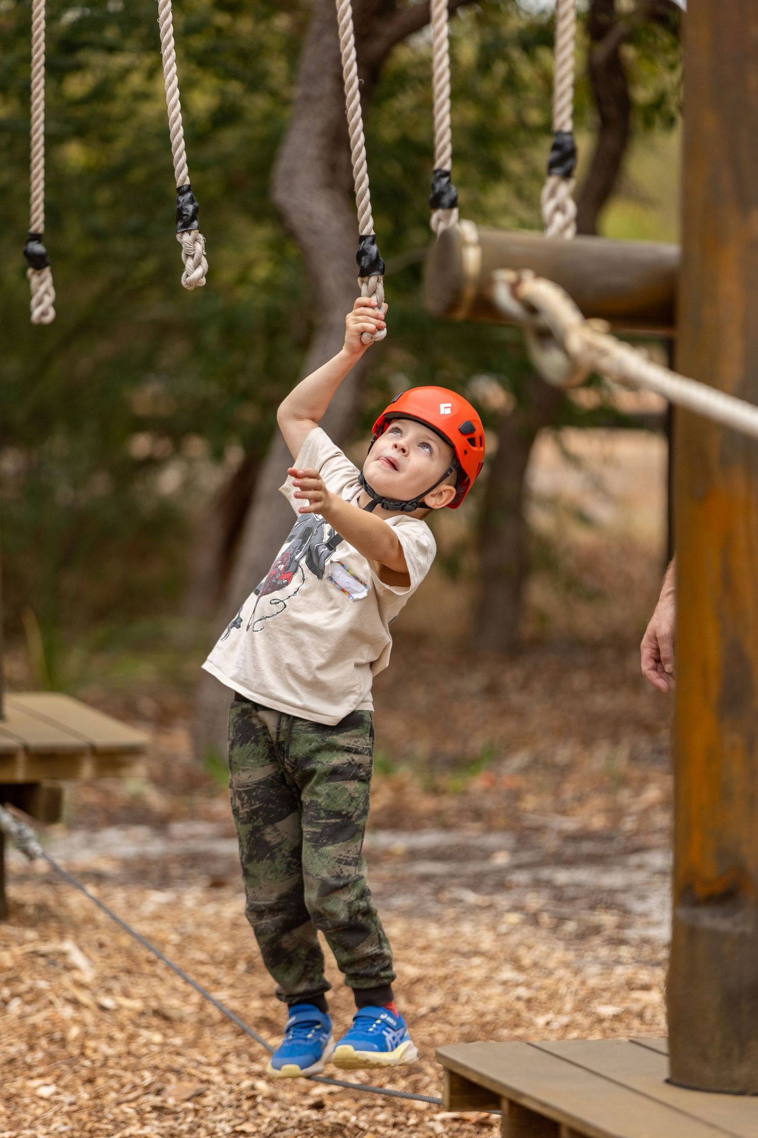 Young adventurer in safety helmet and camo pants holds rope on outdoor climbing course in woodsy setting.