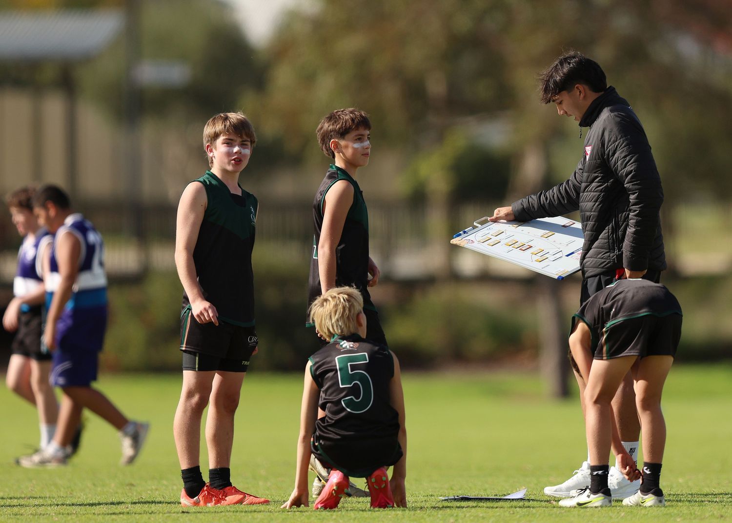 Football players kneel on field during practice session while working with a coach outdoors.