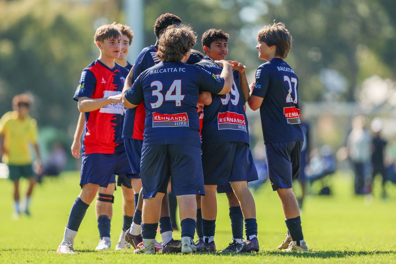 Youth soccer players in navy blue uniforms celebrate together on a green field during a competitive match.