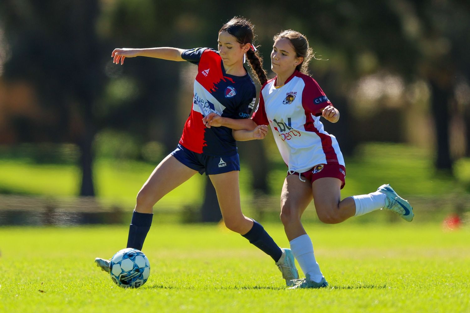 Two female soccer players compete for the ball during an outdoor match, one in red and blue, the other in white and maroon.