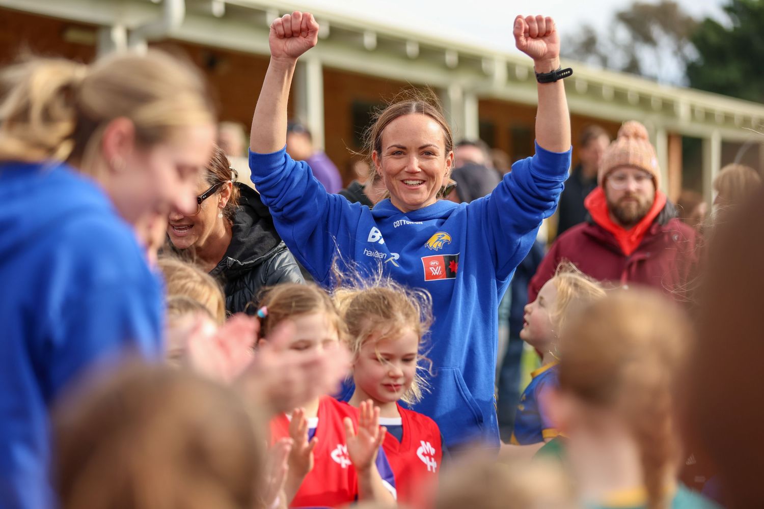 A sports coach in a blue jersey celebrates with arms raised while surrounded by young children at a sports event.