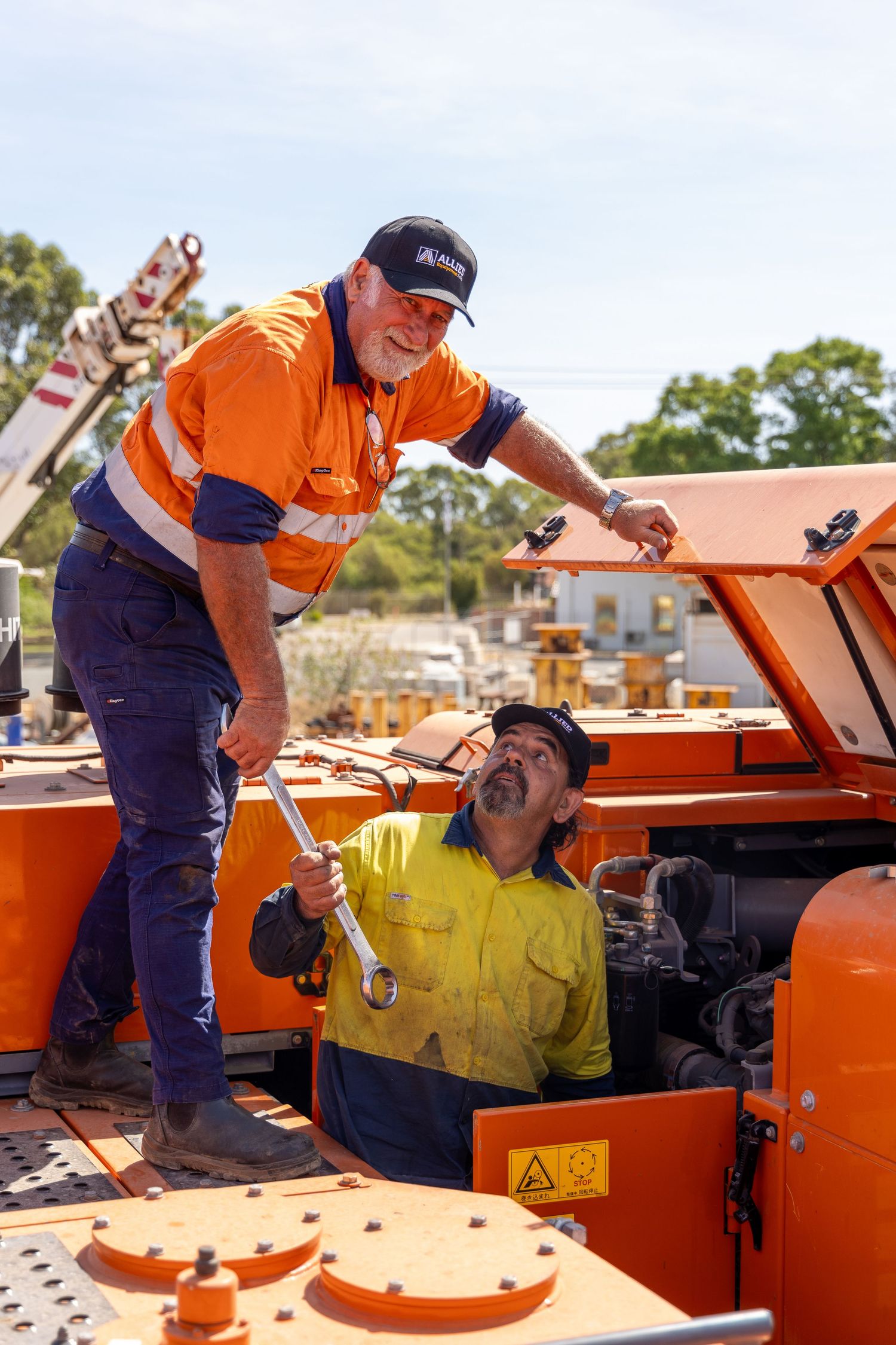 Two workers in high-visibility workwear sharing a laugh while servicing a bright orange industrial machine outdoors.
