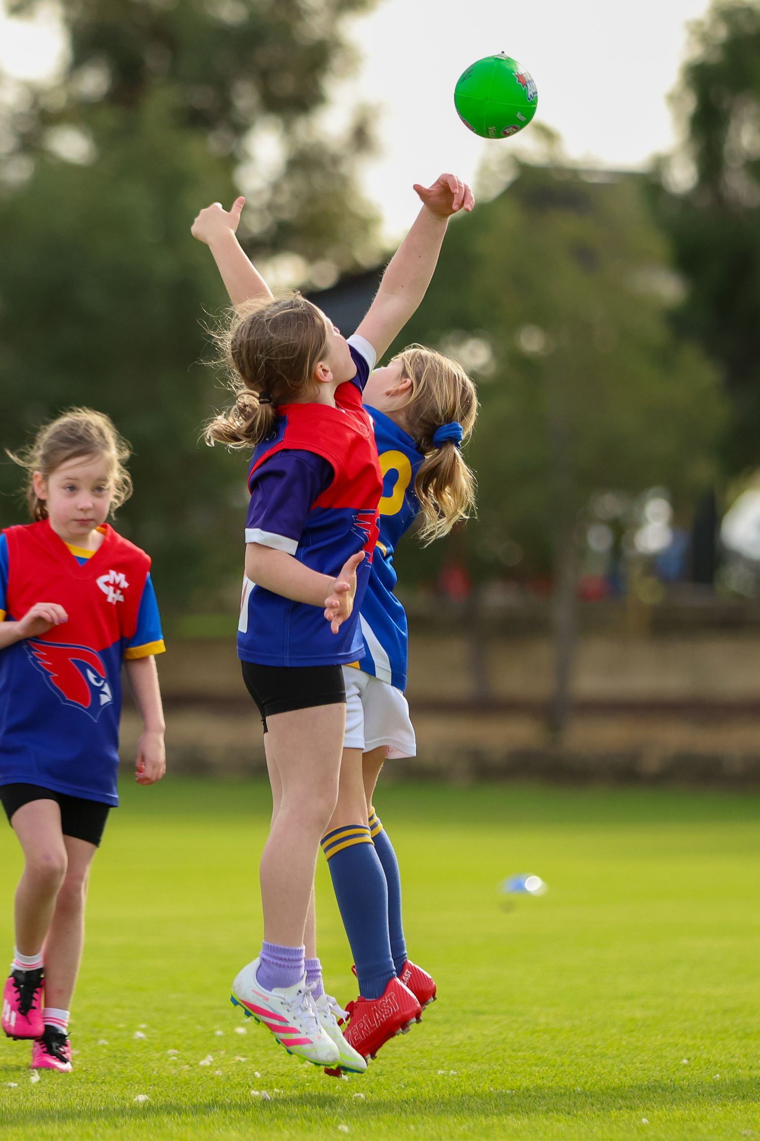 Youth soccer players in red and blue uniforms jump to reach for a green ball on a grass field during a match.