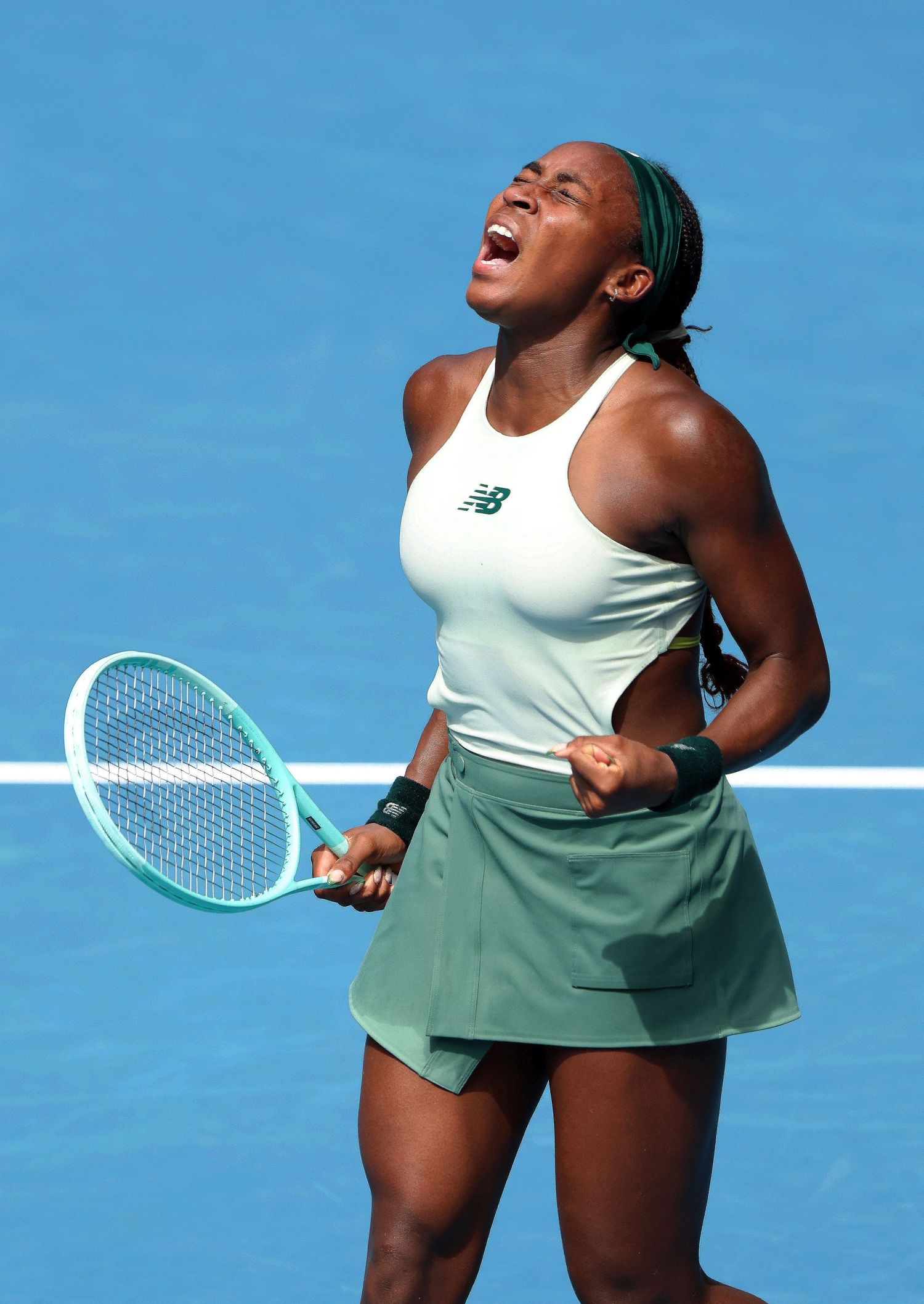 Professional tennis player celebrates emotionally during match against blue sky backdrop wearing white tank top and green skirt.