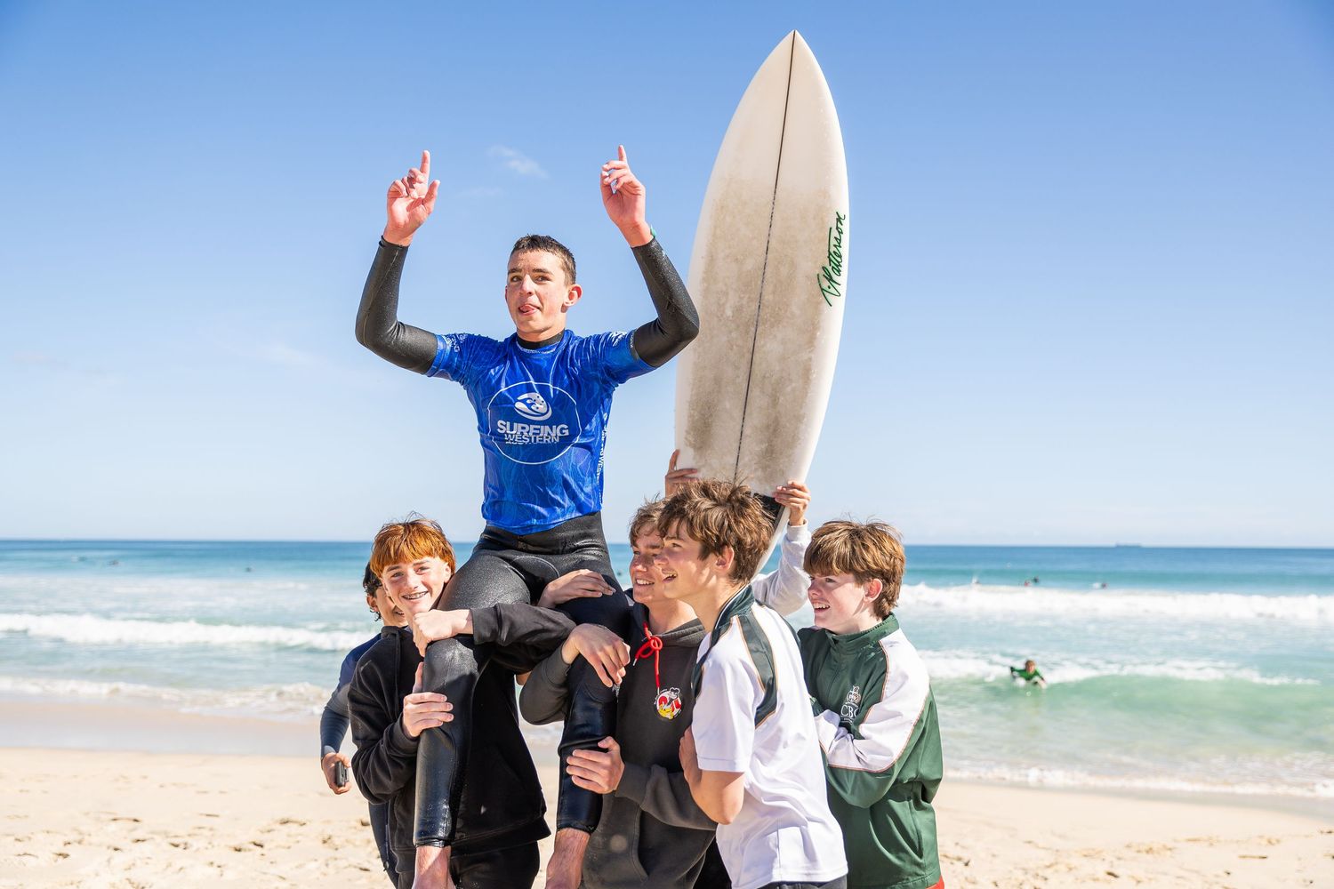Surfers of various ages walk along a sandy beach carrying surfboards on a sunny day with blue ocean waves.