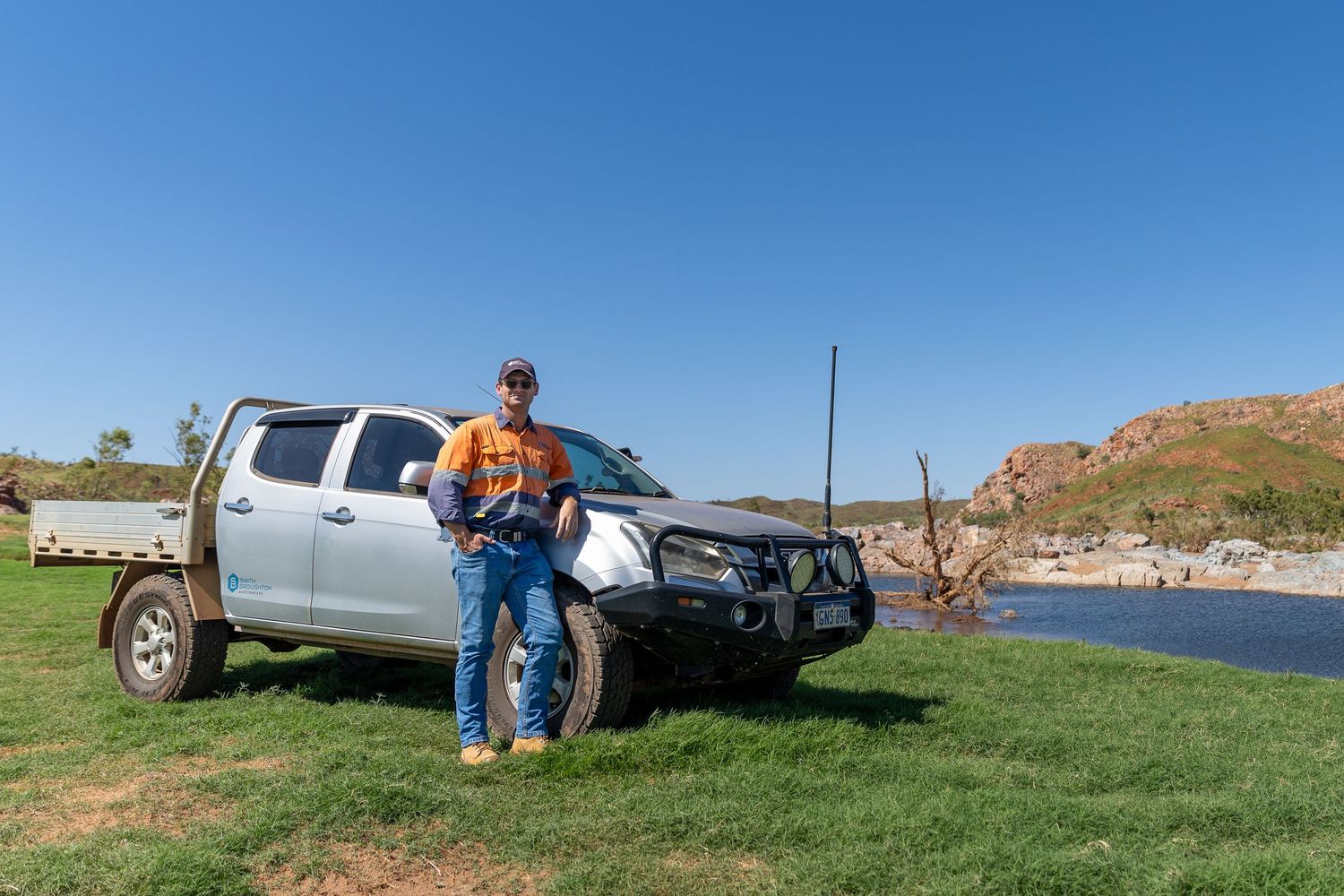 A silver pickup truck with bull bar parked on grass near red cliffs and a lake.