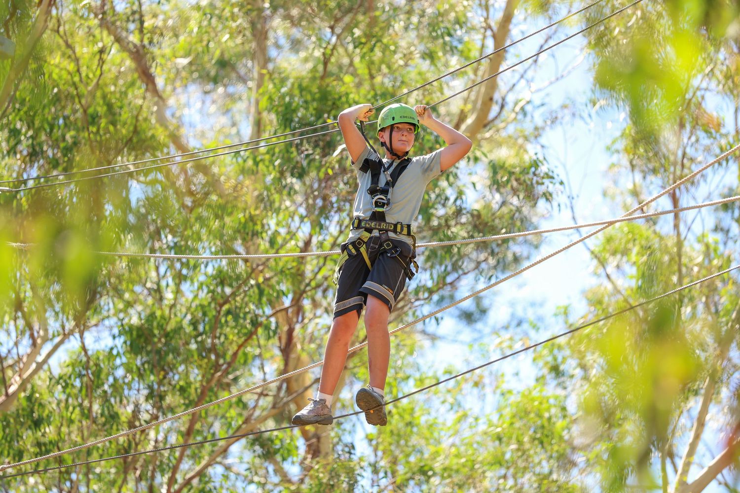 Adventure seeker in safety harness balances on ropes course high up in trees against blue sky and green foliage.