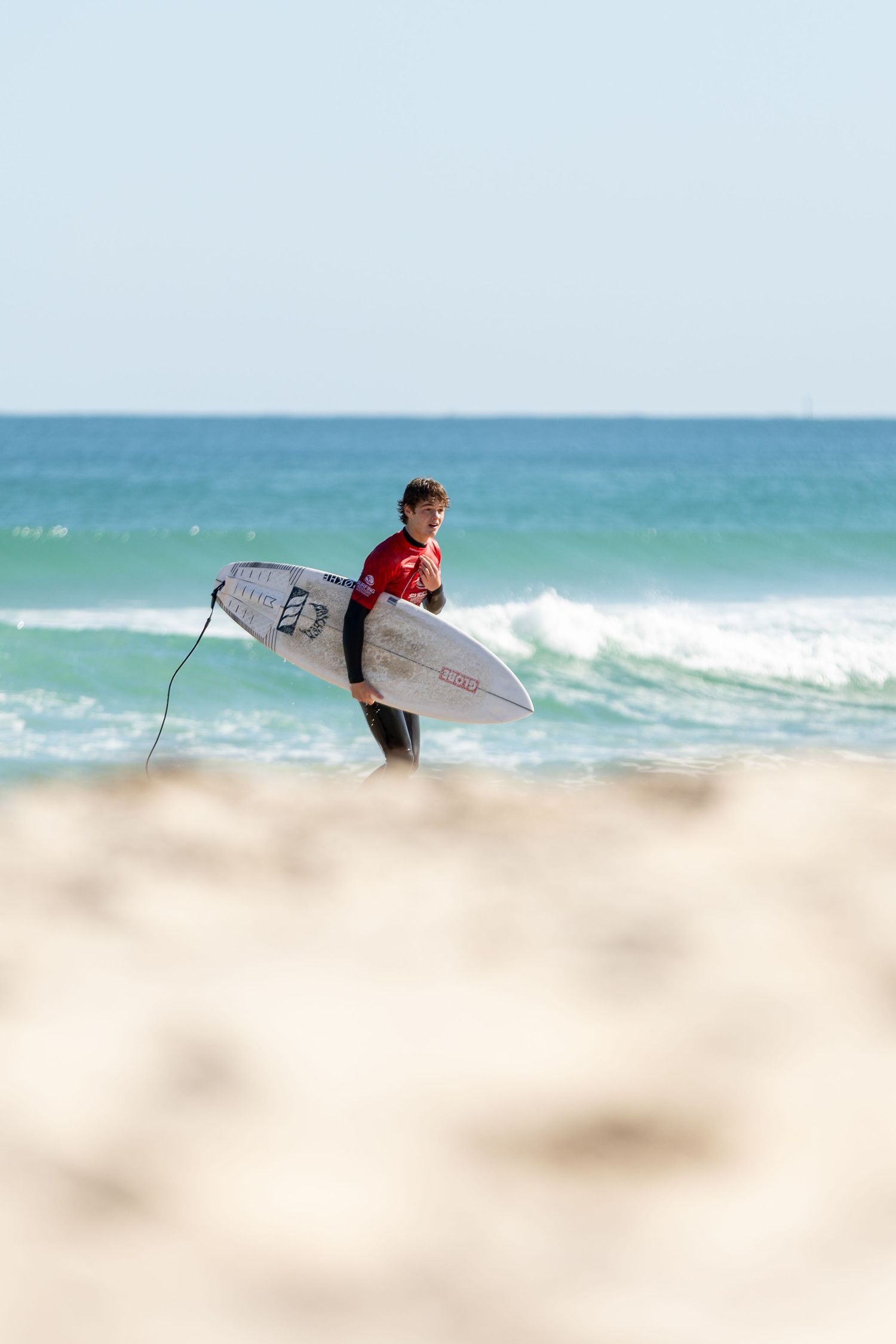 A surfer carrying a white surfboard walks along the beach towards turquoise ocean waves on a sunny day.