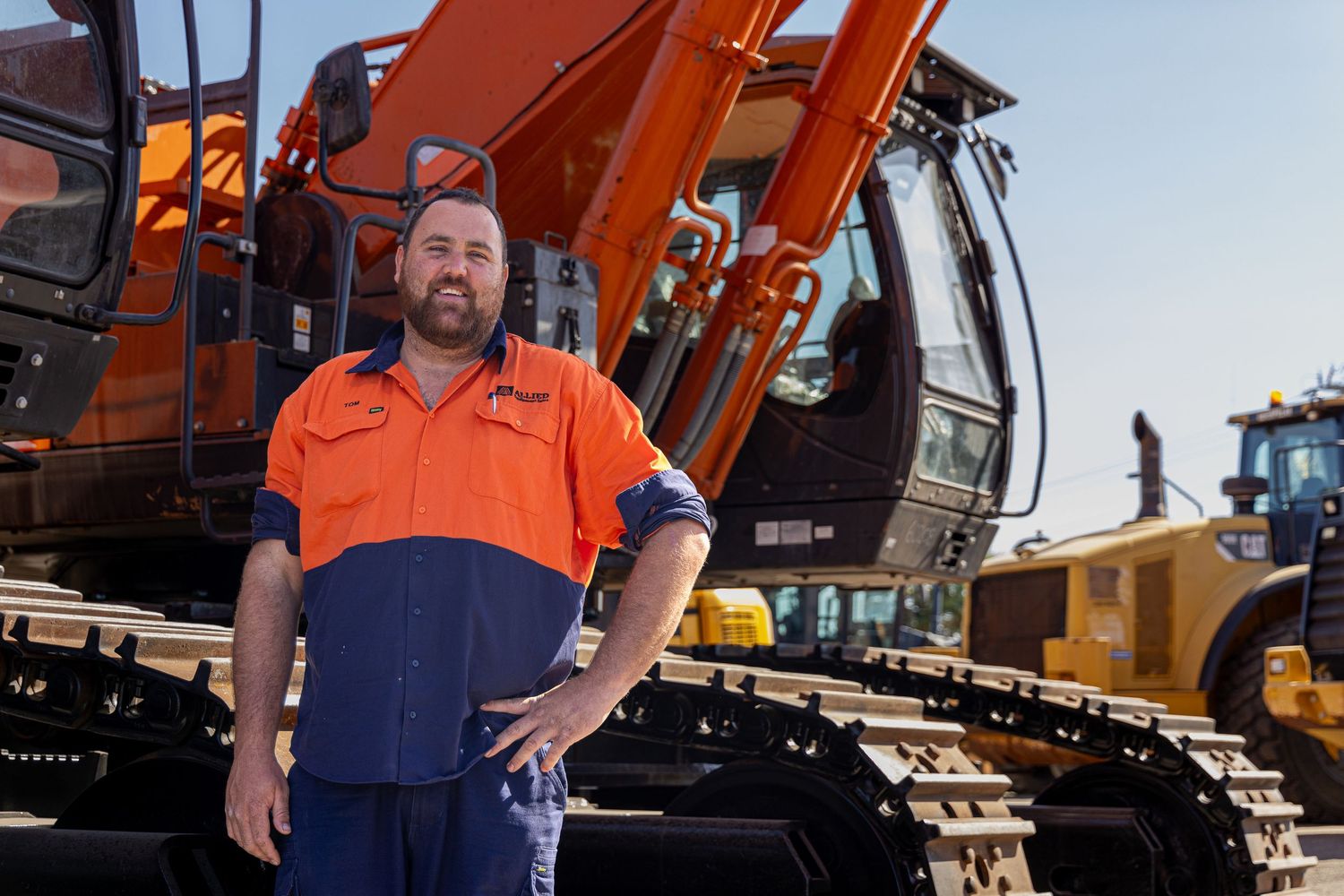 Construction worker in orange safety vest standing in front of large orange excavator machinery and equipment.