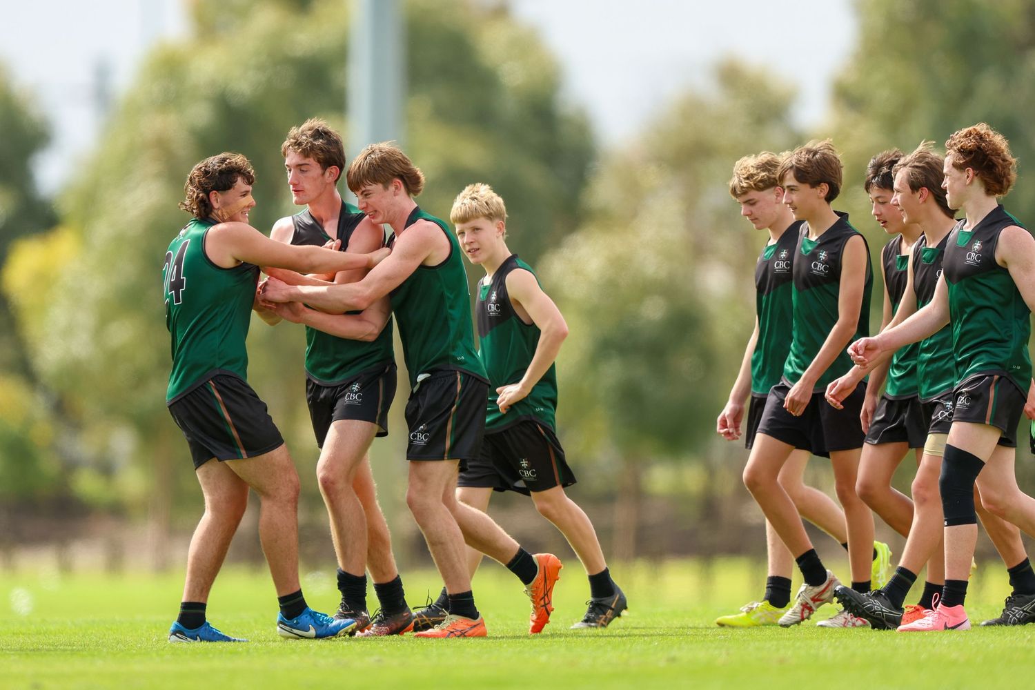 Athletes in green uniforms perform a running drill during team practice on a sunny grass field.