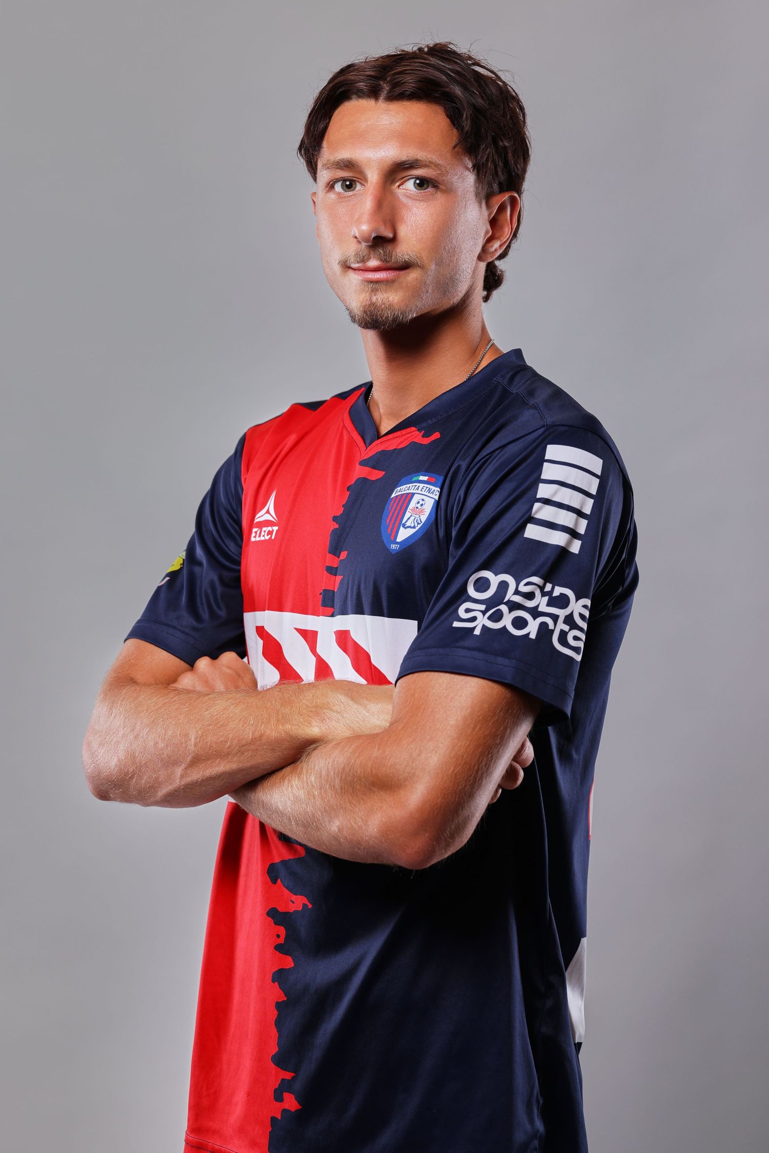 Professional athlete poses in red and navy blue sports jersey with crossed arms against gray background.