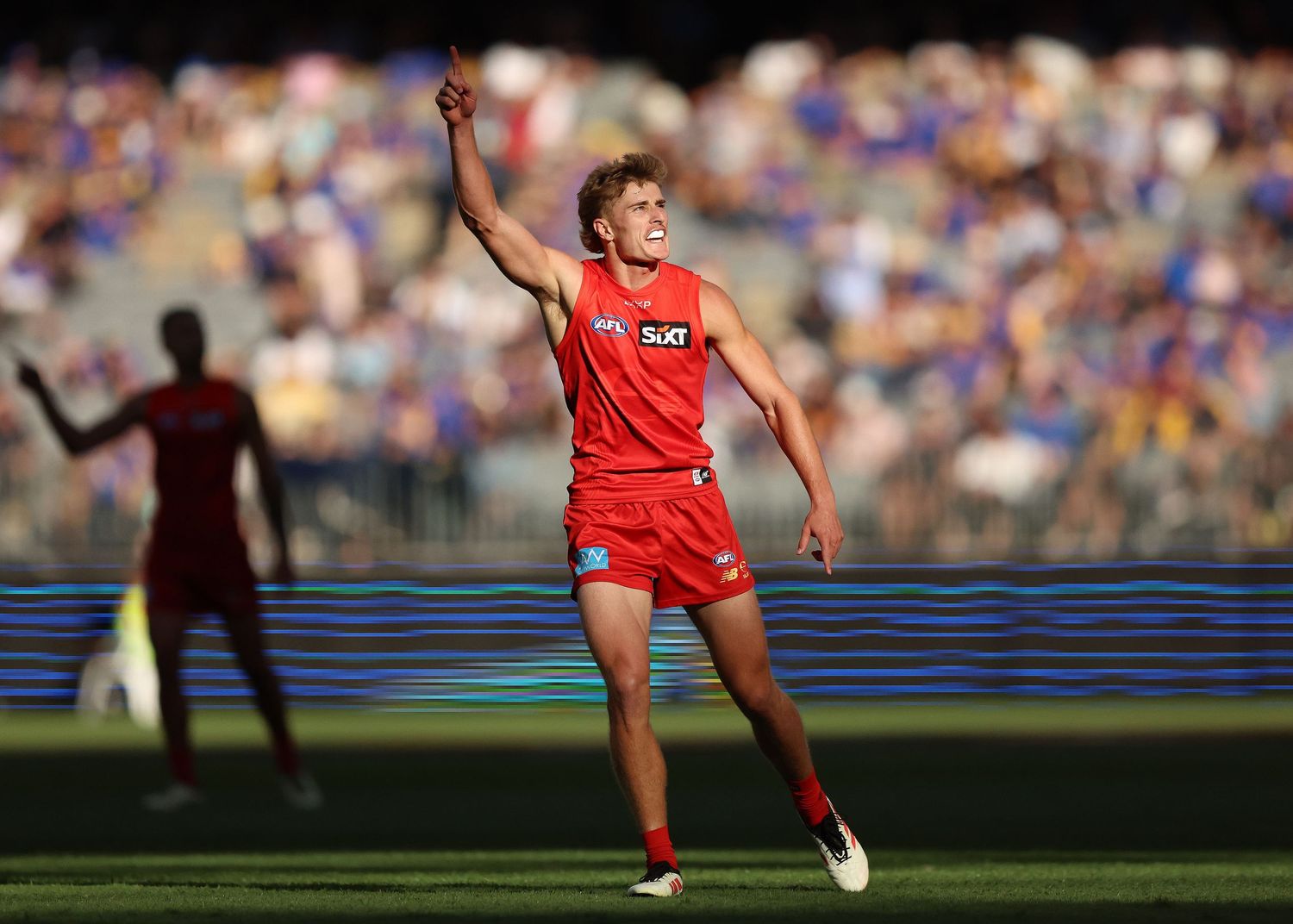 AFL player celebrating with raised arm in red uniform against stadium crowd.