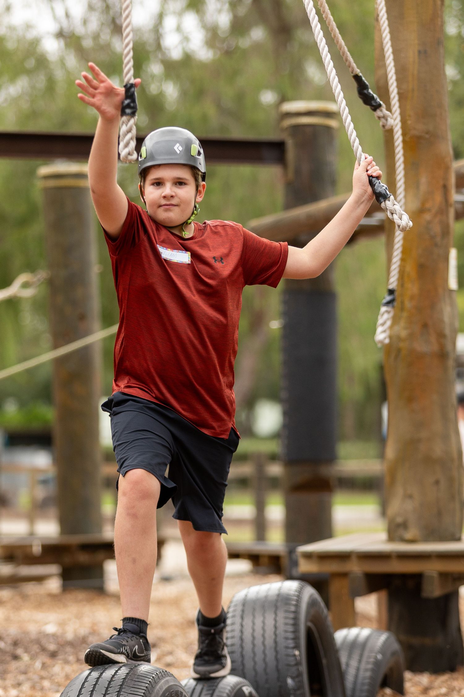 Child in red shirt and safety helmet balancing on tires while holding ropes at an adventure course.