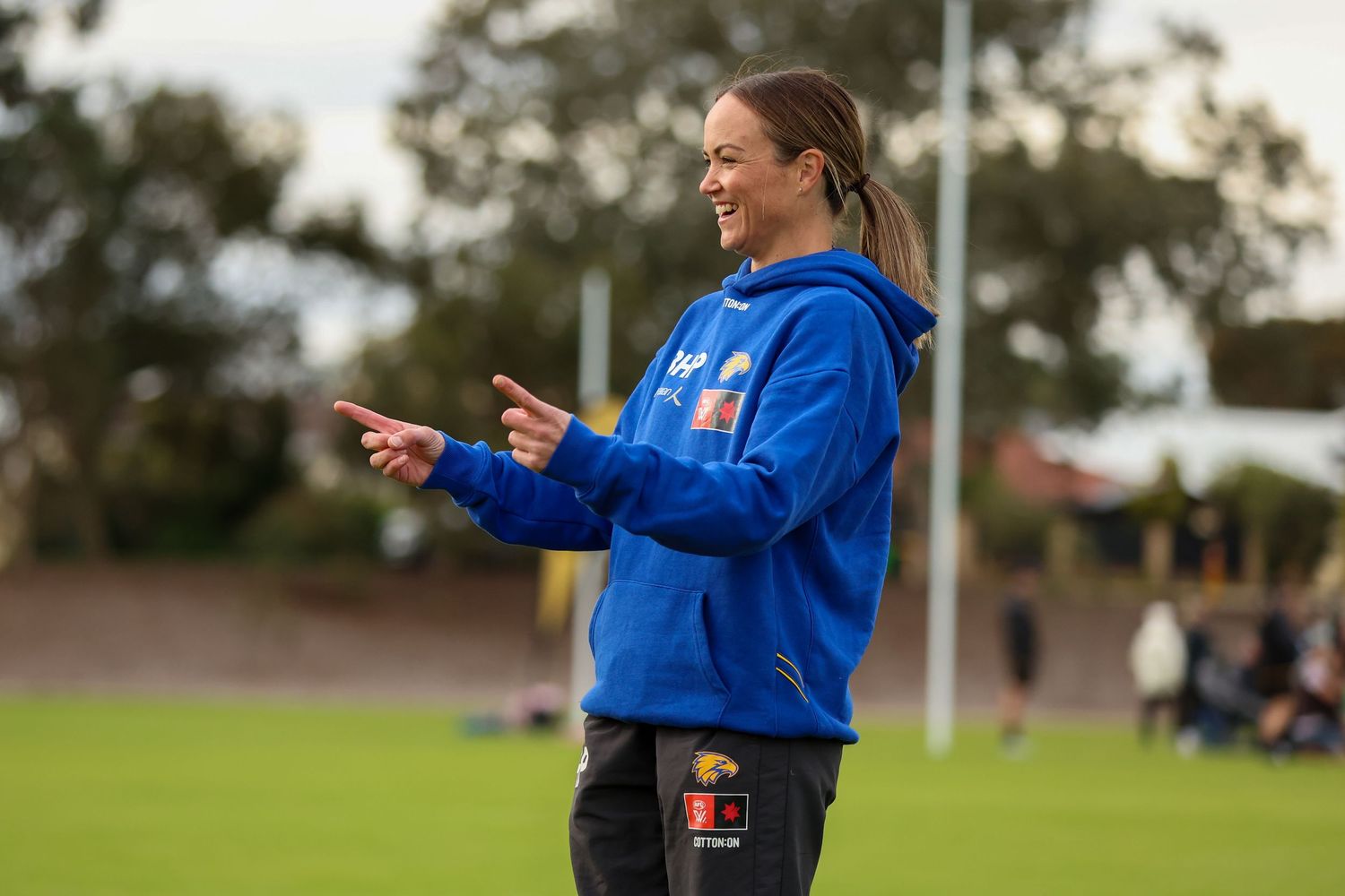 A coach in a blue hoodie gestures with both hands while directing from the sideline of a sports field.