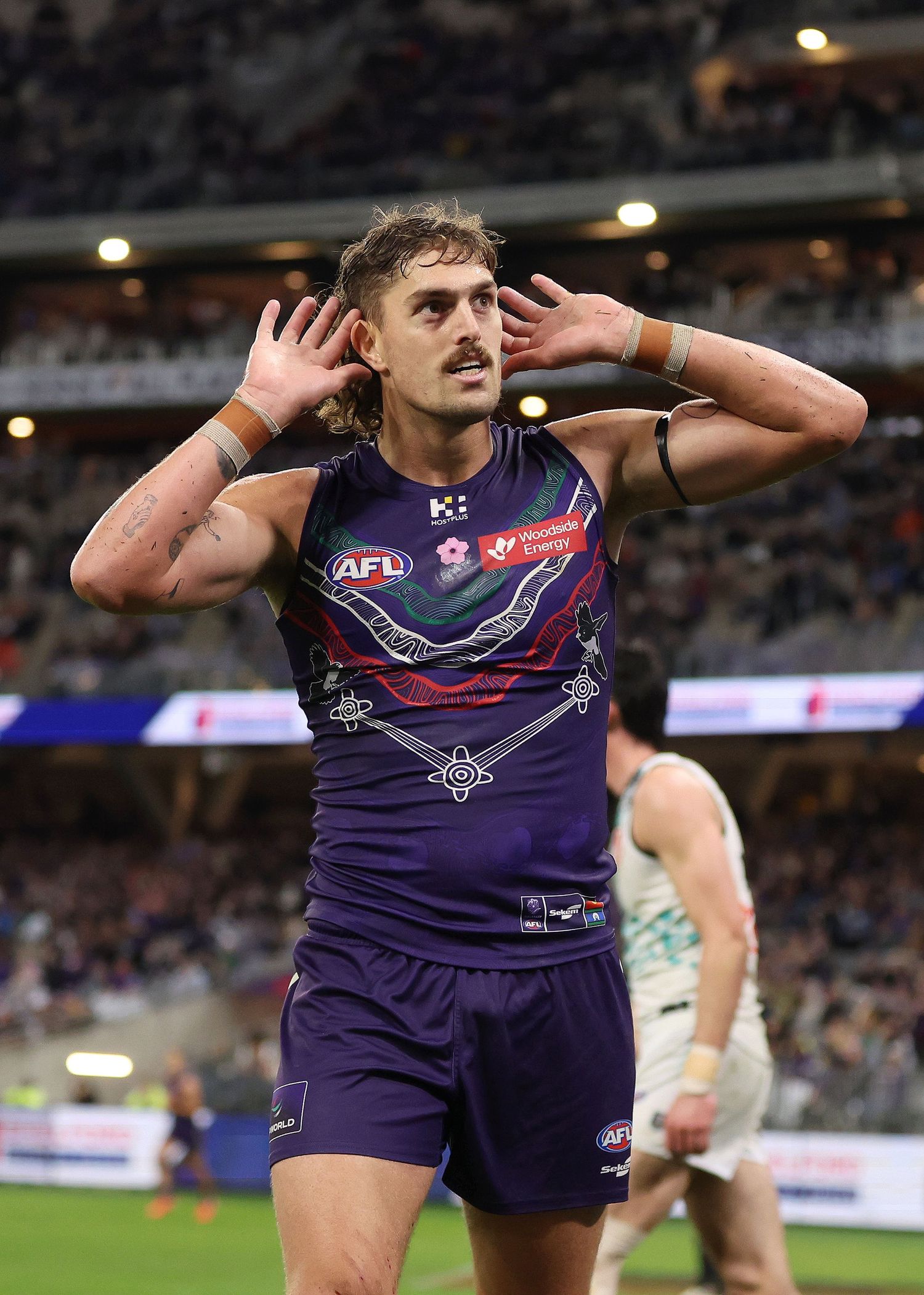 Australian Rules Football player in purple uniform celebrates goal with hands raised in salute during match.