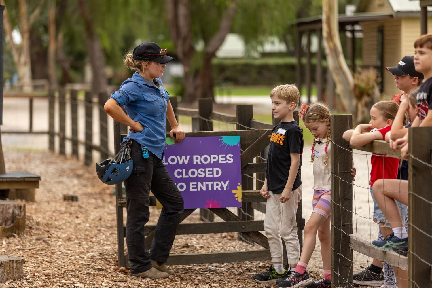 Children and visitors gather around a purple 'Low Ropes Closed' sign at an outdoor recreation area.
