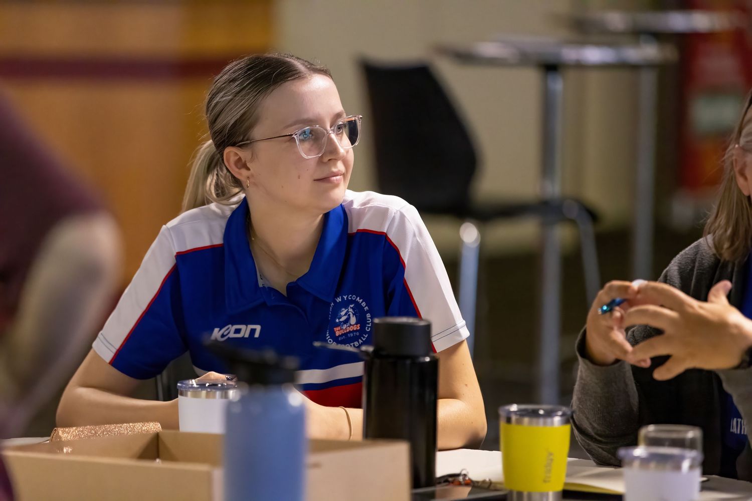 A student wearing glasses sits at a table while working on their studies in a classroom environment.