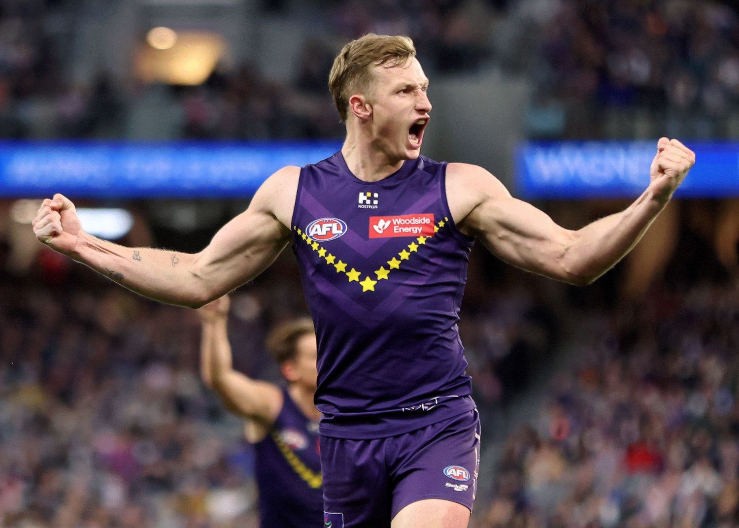 AFL player in purple Fremantle Dockers jersey celebrates with arms outstretched during match at stadium.