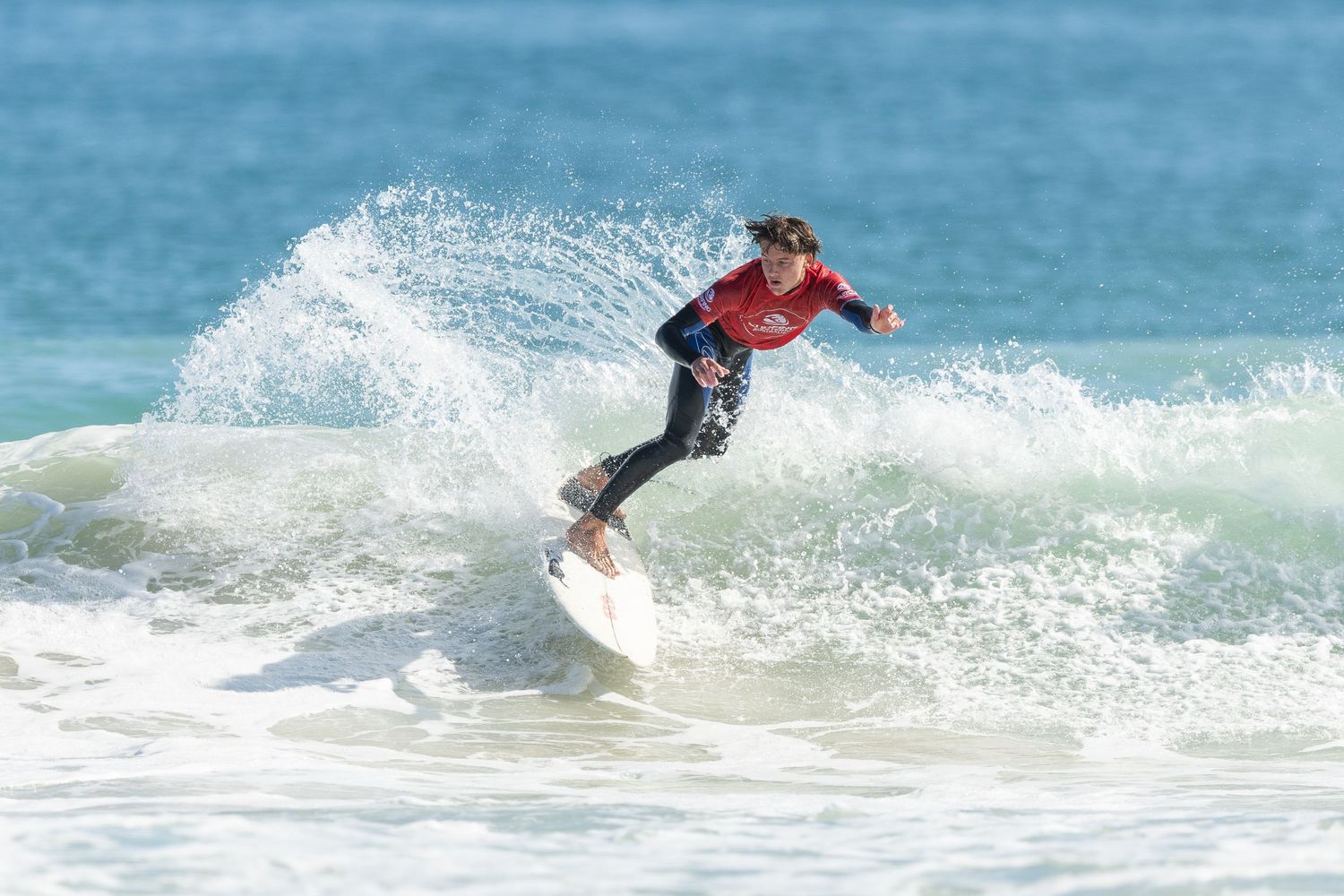 A surfer performs a dynamic aerial maneuver off a wave against a bright blue ocean backdrop.