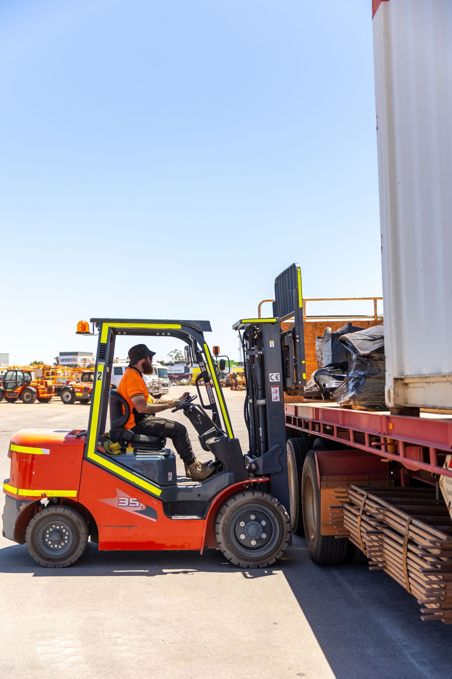 Operator using orange forklift to load cargo onto flatbed truck trailer in industrial yard under blue sky.