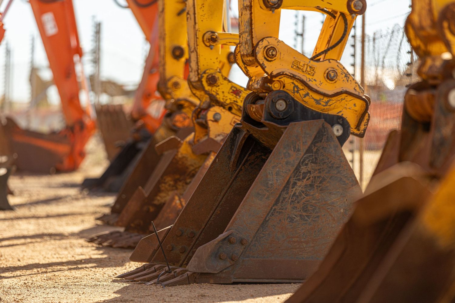 A row of rusty excavator buckets cast shadows in warm sunlight at a construction equipment yard.