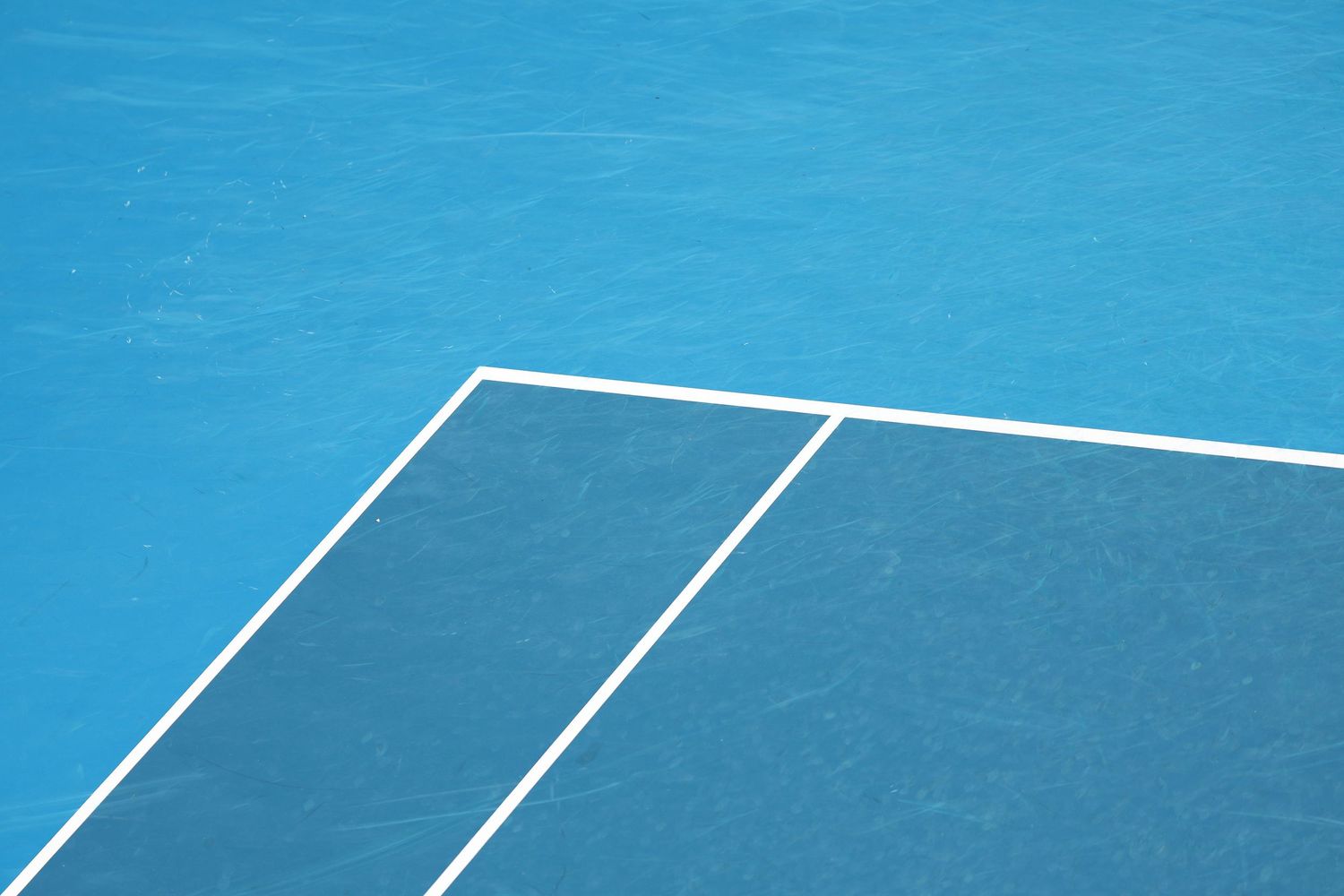 White boundary lines intersect on a bright blue tennis court surface creating geometric angles and shapes.