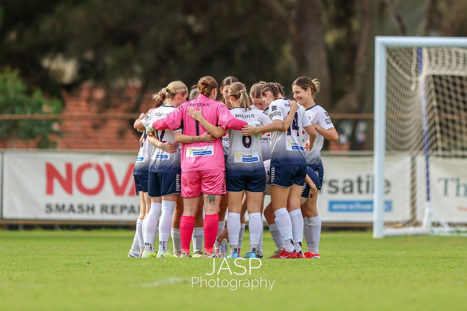 Women's soccer team huddles together on green field during a match, wearing navy and pink uniforms.