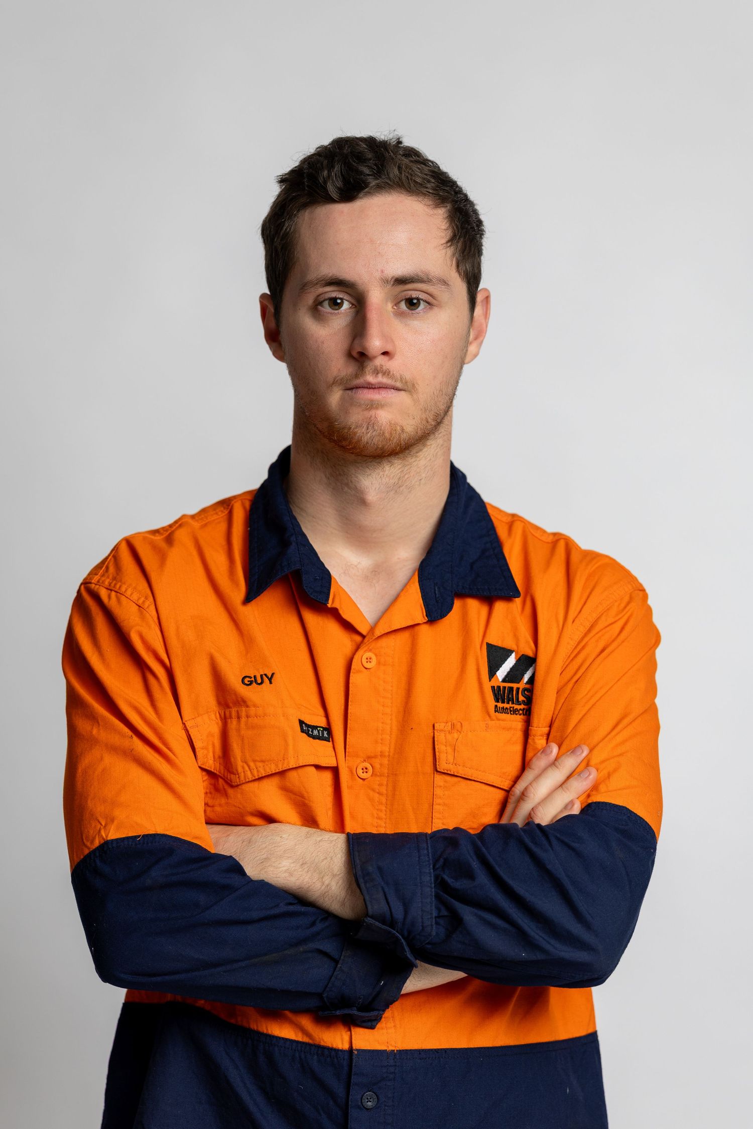 Portrait of worker wearing orange and navy hi-vis workwear uniform with crossed arms against gray background.