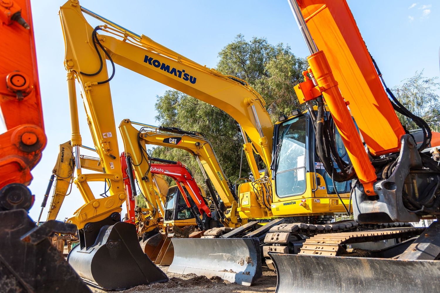 Row of yellow and orange hydraulic excavators lined up at a construction equipment yard with trees in background.