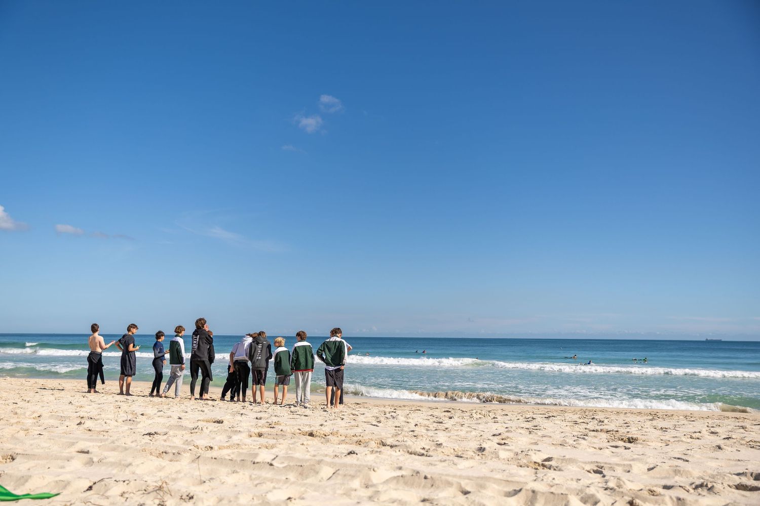 Group of people standing along a scenic beach shoreline with clear blue sky and ocean waves in the background.